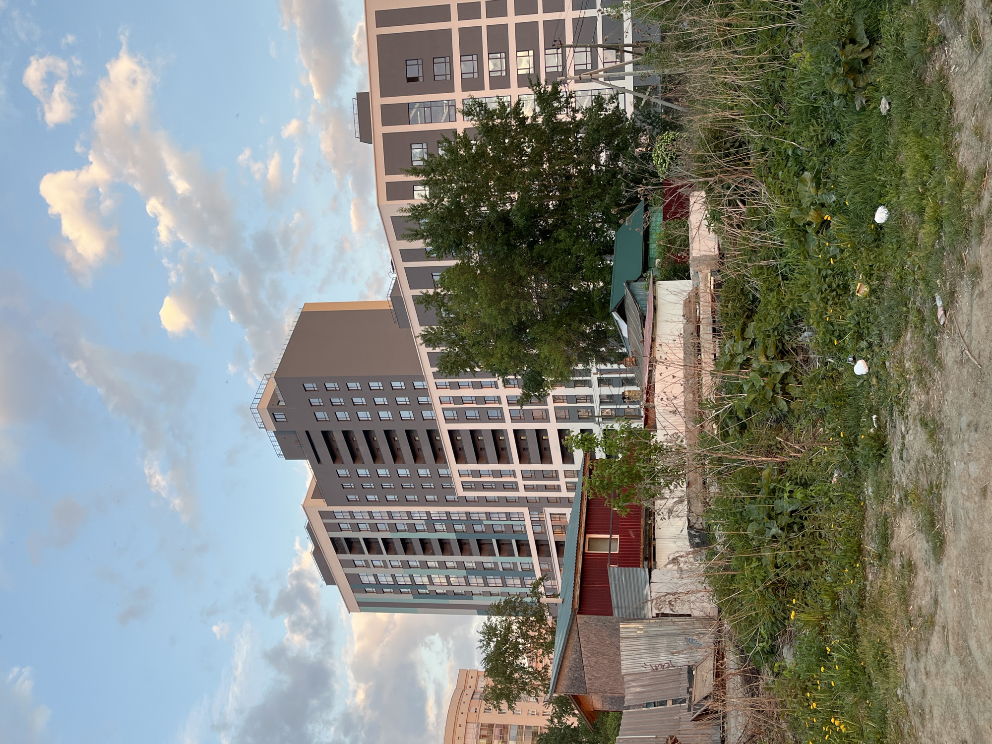 The image depicts a juxtaposition of modern and dilapidated structures in Yekaterinburg, Russia. In the background, tall, multi-story apartment buildings dominate the skyline, with a neutral gray and white color scheme and repetitive window patterns. A large green tree partially obscures one of the buildings.
In the foreground, a collection of small, run-down structures is visible. These appear to be makeshift or poorly maintained buildings, with corrugated metal and weathered wood comprising their facades. Overgrown vegetation and debris are scattered around the base of these structures and along a dirt path, indicating neglect and disrepair.
The sky is filled with scattered clouds, and the overall lighting suggests it is either early morning or late afternoon. There are no people visible in the frame. The image captures a stark contrast between the modern, urban development and the remnants of older, neglected structures within the same urban environment.