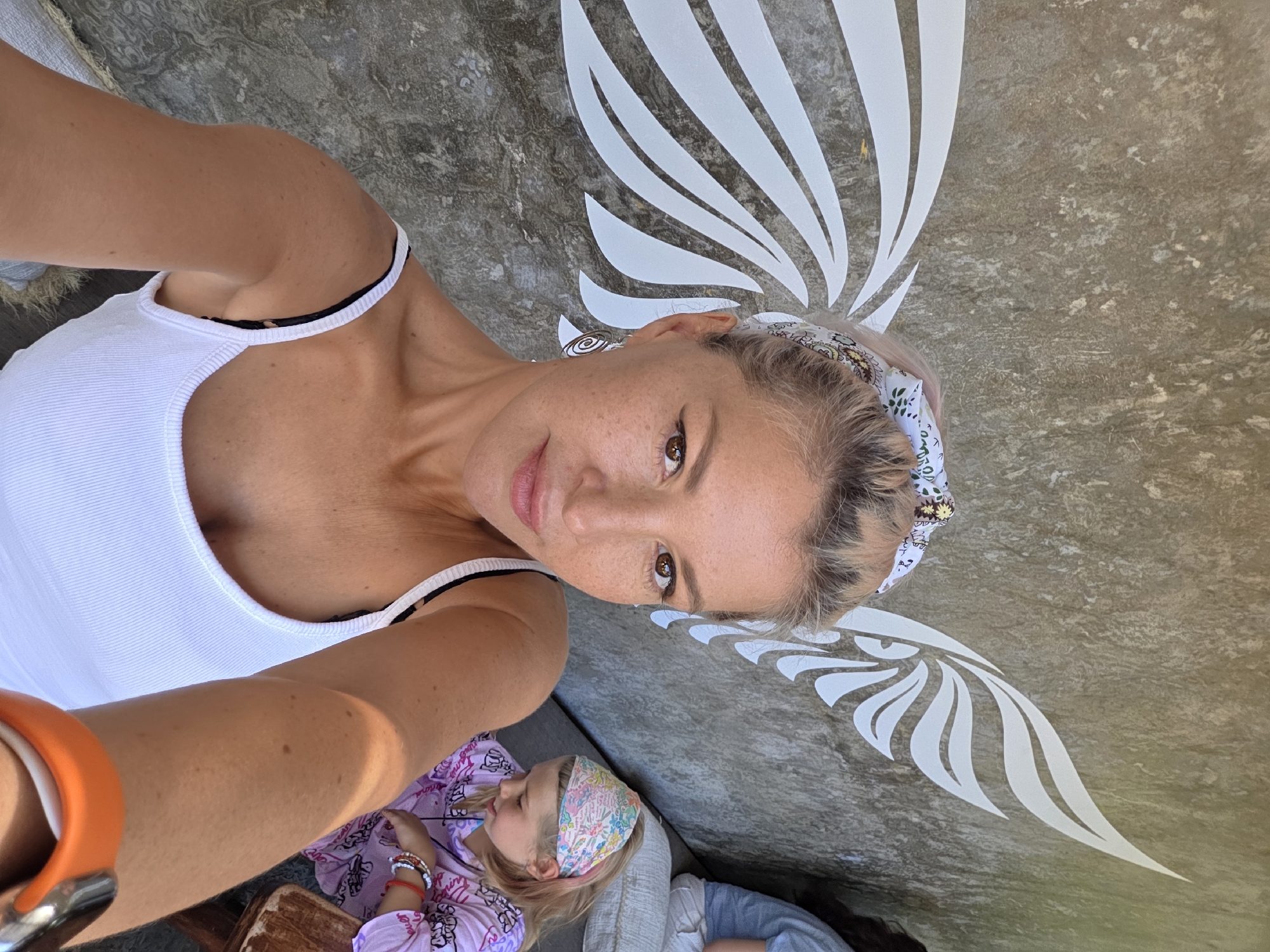The image is a selfie taken in North Kuta, Indonesia. A woman with blonde hair and a white tank top is holding the camera. She has an orange watch on her wrist. A child is lying down in the lower part of the frame, wearing a patterned headband and a pink shirt. Behind the woman are white wing decorations on a textured wall.