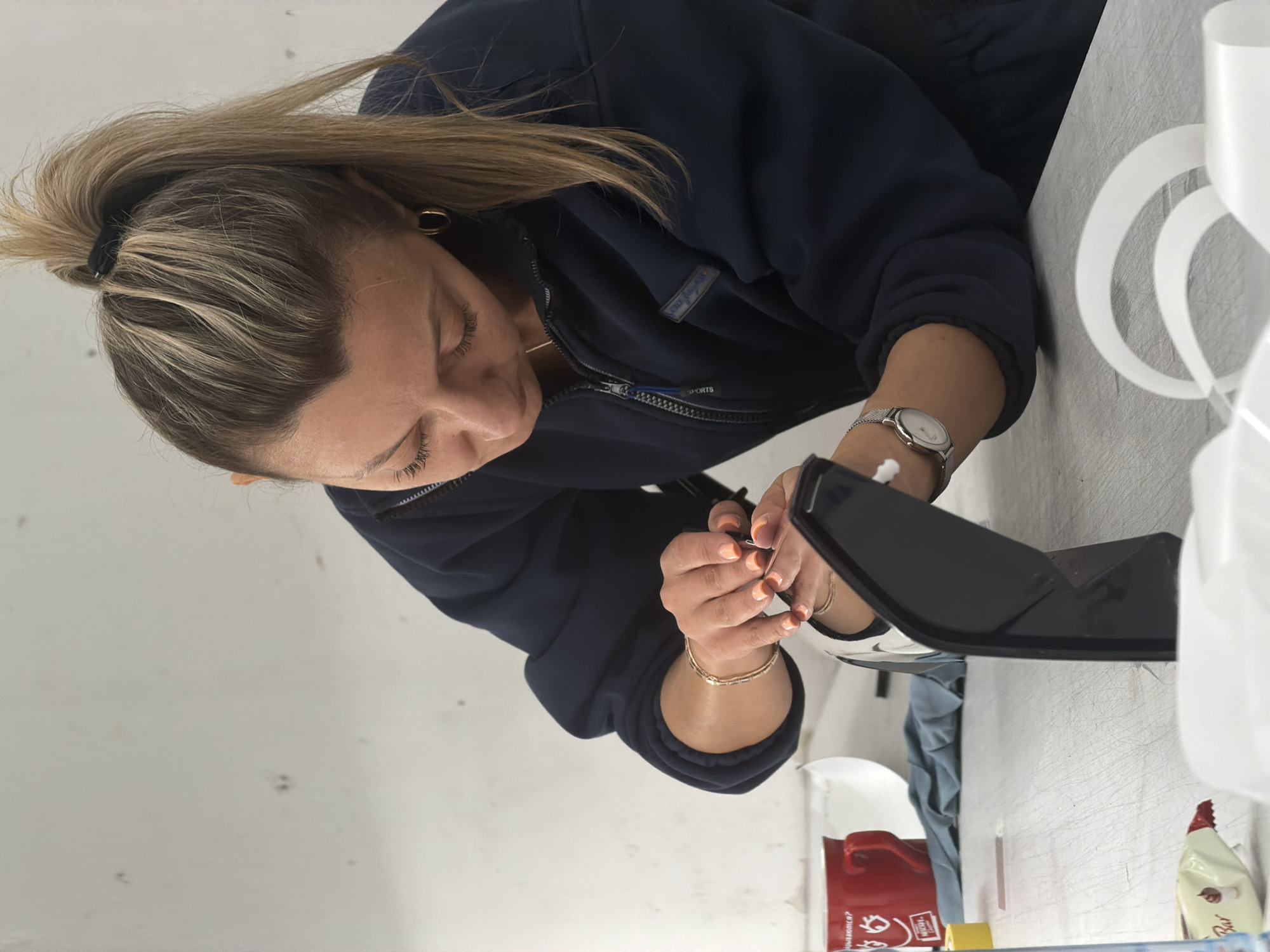 A woman with blond hair pulled back in a ponytail, wearing a navy blue zip-up jacket and gold jewelry, is sitting at a table. She is holding a small black object with her hands, and her attention appears focused on it. A watch is visible on her left wrist. The table is covered with various items, including what appears to be packaging materials, a red container, a white dispenser, and a tube of lotion or cream. Based on the presence of Cyrillic text on the red container, the location may be in a Russian-speaking region. While Krasnoyarsk, Russia, cannot be confirmed based solely on the image, the presence of Cyrillic text supports that this is taking place in Russia.