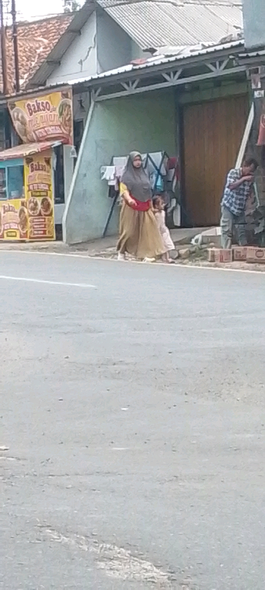 In Cilaku, Indonesia, on a street next to a building, a woman wearing a grey hijab and a tan skirt with a red accent is walking while holding the hand of a young girl in a light-colored dress. To their right, a man in a blue and white plaid shirt and grey pants is leaning against a wall, with his hand near his mouth. A food stall with yellow signage advertising "Bakso" (meatball soup) is visible to the left. The environment appears to be a commercial area with a road in the foreground and a building with a corrugated metal roof in the background.