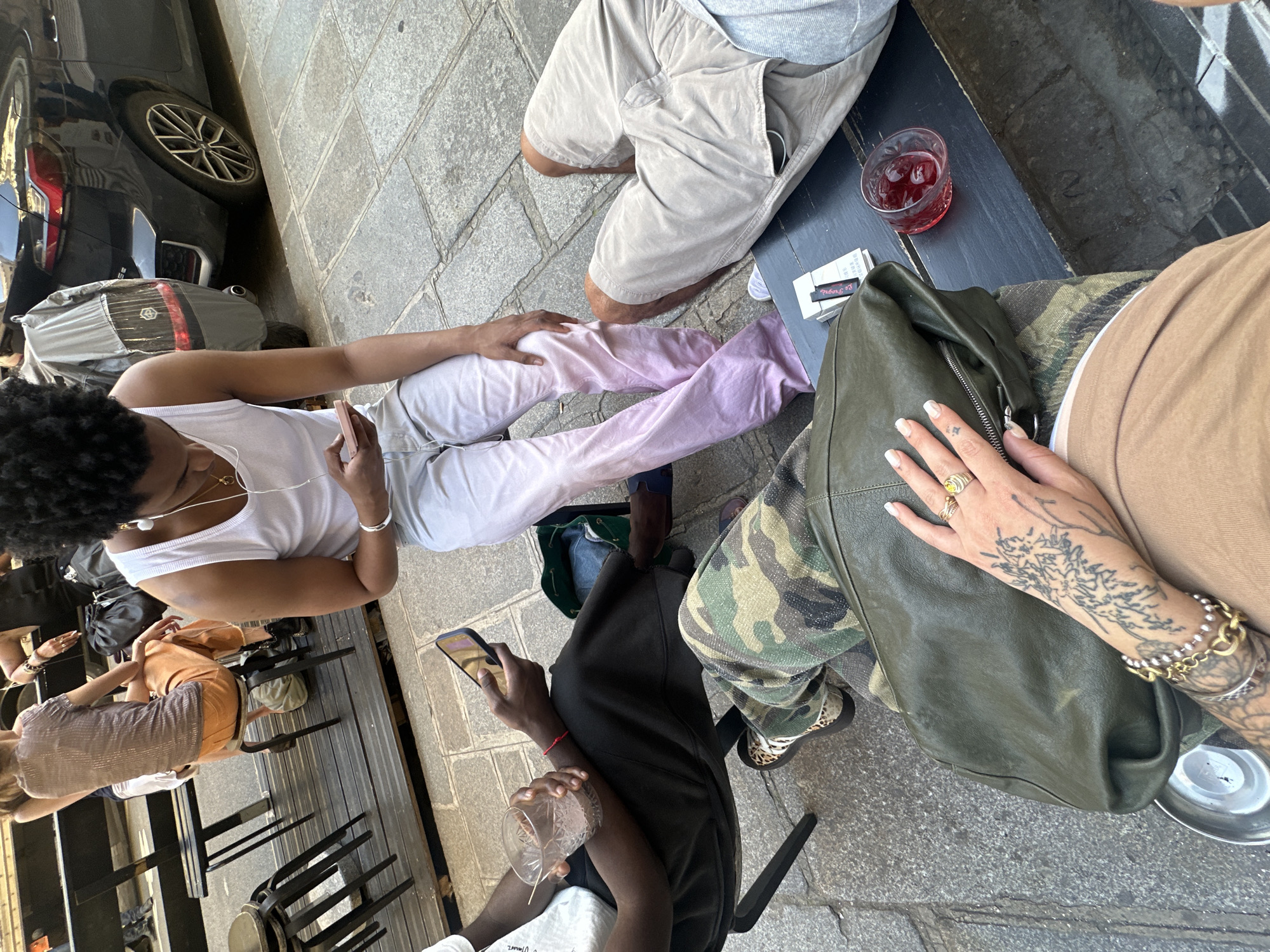 The scene is a street-level view, likely in Paris, France, based on the architectural details and ambiance. The image appears to be taken from an elevated perspective looking down at a small outdoor gathering. The foreground of the image consists of people seated around a table in an outdoor cafe or bar setting. There are several people visible, mostly from the torso down, and some are holding phones and drinks. There's a table with glasses, a pack of cigarettes, and what appears to be a drink with ice. Some have legs are crossed and are wearing different types of pants and shorts, and their footwear. The background includes the edge of what appears to be a black car parked on the side of the street, along with hints of building facades and other cafe chairs. The environment gives off the feel of a casual outdoor social gathering.