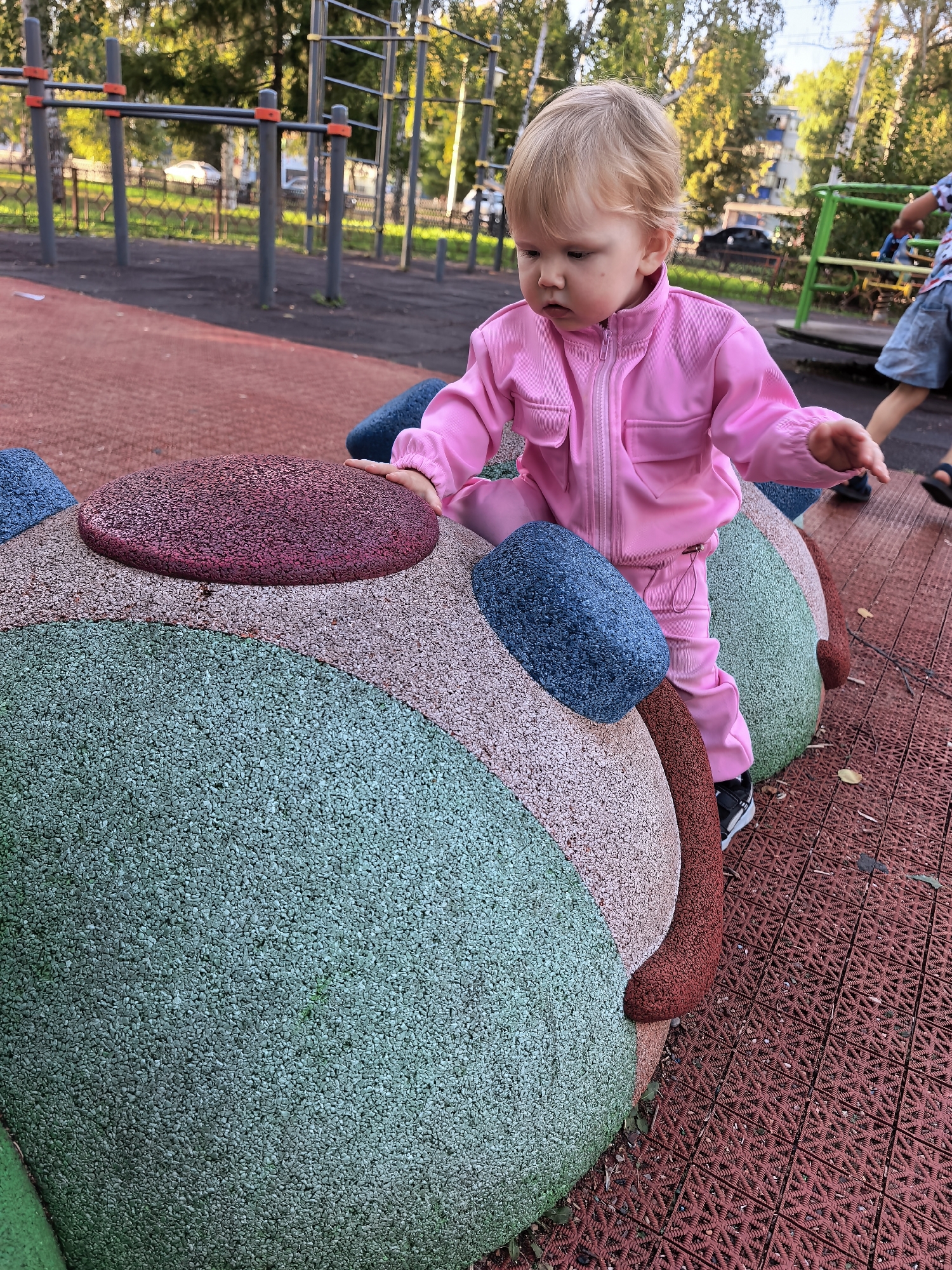A young Caucasian child, likely a toddler, with blonde hair is seated on a colorful, textured playground structure resembling a large caterpillar or bean. The child is wearing a pink tracksuit and dark sneakers. The structure is composed of rounded segments in various muted colors including green, light brown, dark red, and blue. The playground features a red rubberized surface with a textured pattern, and in the background, there are elements of a park or outdoor sports facility with metal bars, trees, and a building. The overall scene is outdoors during daylight.