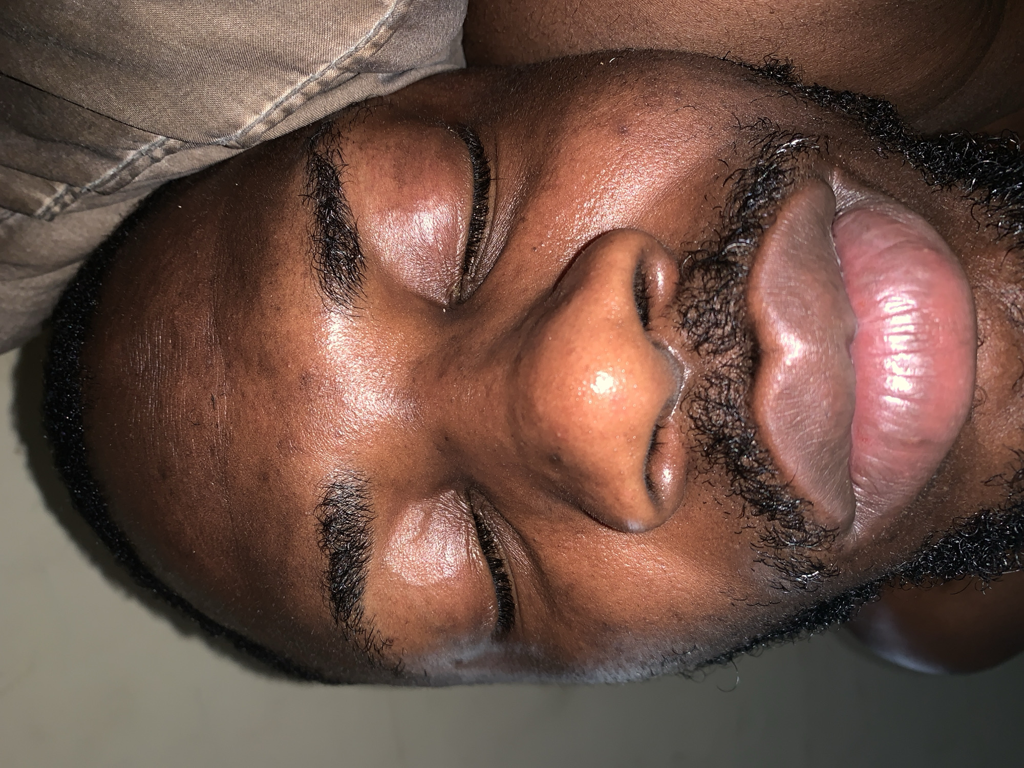 A close-up, upside-down view of a dark-skinned male face. The individual is lying down with eyes closed. The mouth is slightly open. There is stubble and a sparse mustache present. Part of a brown fabric is visible in the upper left corner. The setting is possibly indoors, based on the lighting. No discernible features indicate a location in Rafin Par, Nigeria.