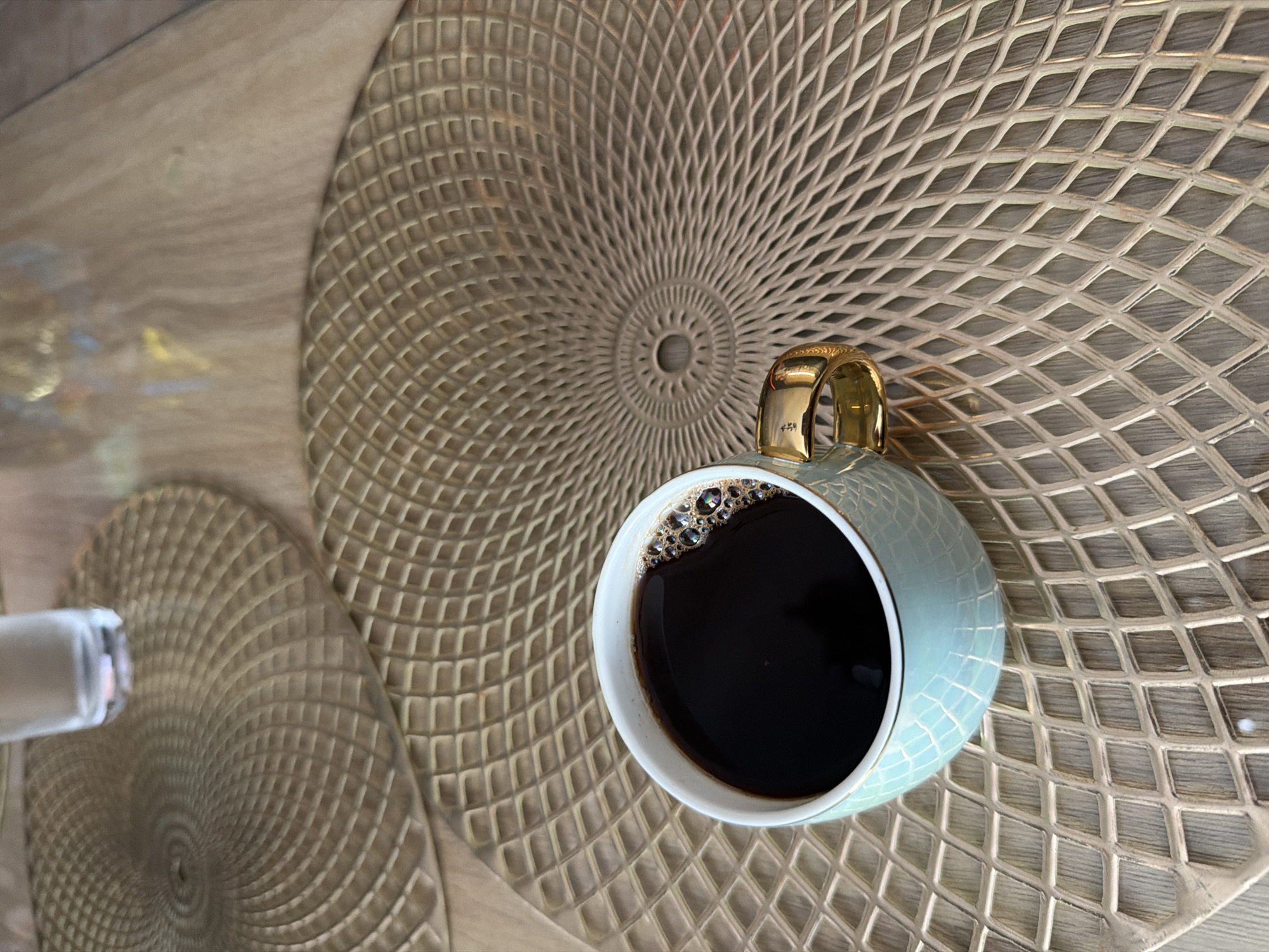 A close-up, eye-level shot shows a light blue, textured coffee cup with a gold handle, filled with dark coffee, resting on a gold, circular placemat with a diamond pattern. The placemat is on a light wood-grain surface, which appears to be a table. Another similar placemat is partially visible in the upper left quadrant of the image, with a glass of water on it. The scene is well-lit, with reflections visible on the coffee and the glass. The overall setting suggests a breakfast or coffee break in a modern, possibly coastal, environment, such as a cafe or restaurant in Rafailovići, Montenegro.