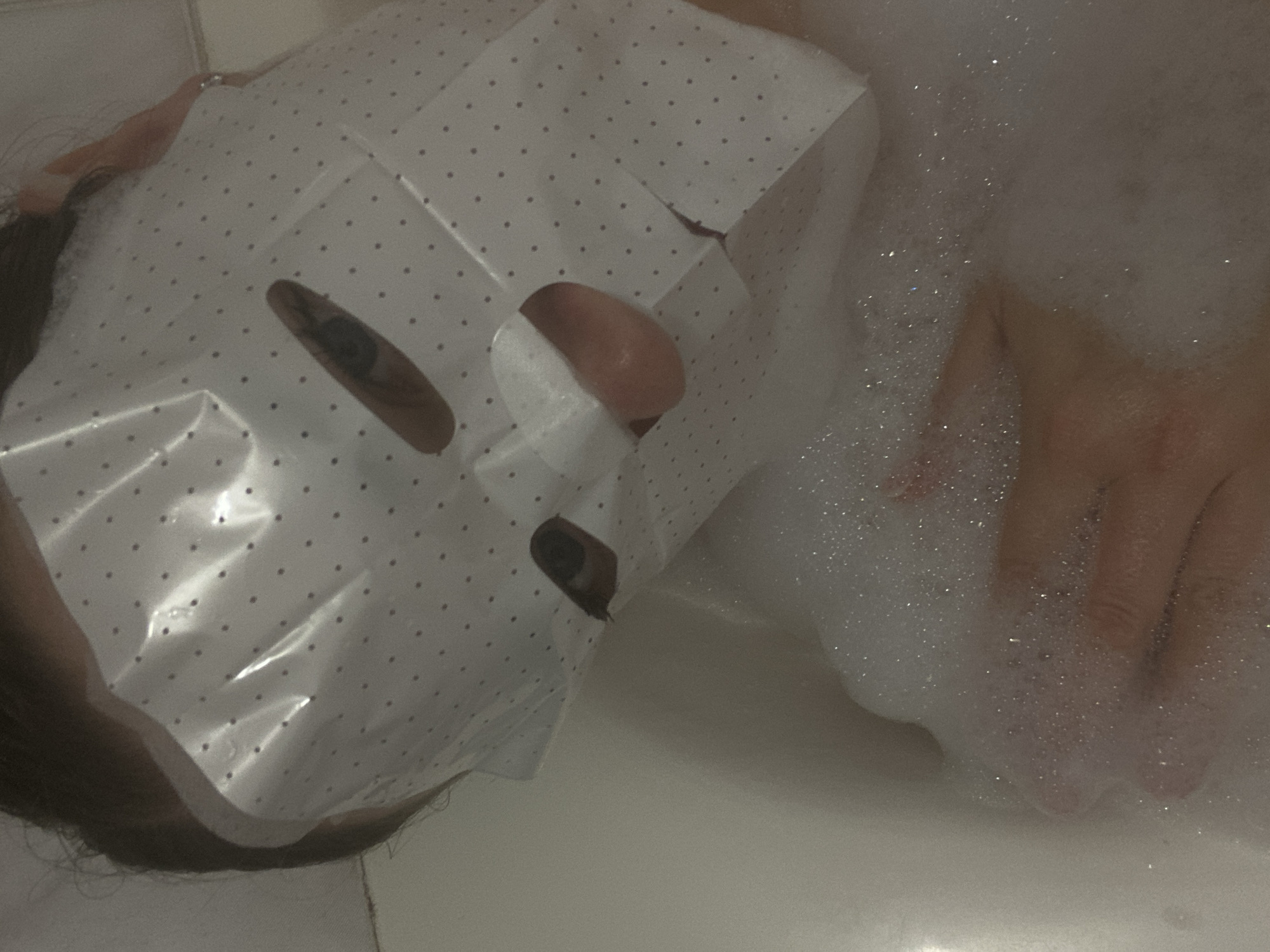 The image displays an individual submerged in a white bathtub filled with white foam. The person's face is covered by a white facial sheet mask patterned with small dark polka dots, featuring visible cutouts revealing parts of both eyes, specifically the irises and pupils, and the upper lip/philtrum area. Dark hair is visible along the upper edge of the mask. One hand is partially visible beneath the surface of the bath foam. The setting is an indoor bathroom, indicated by the presence of a bathtub. No specific visual information is present to identify a precise location within the city of Sochi, Russia.