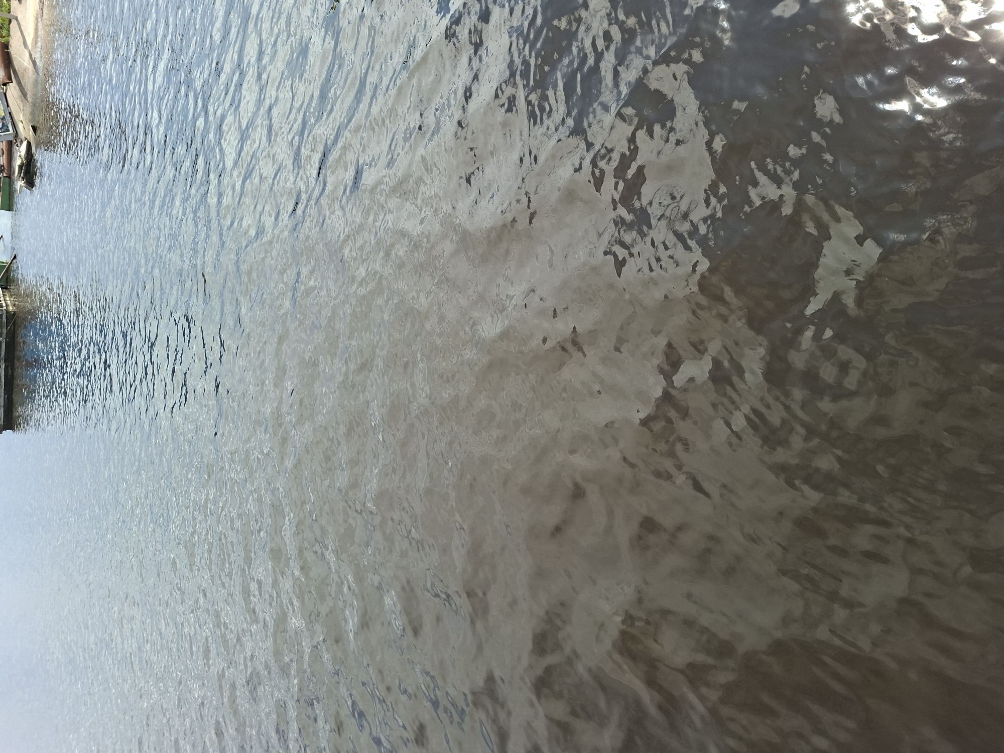 The image shows a body of water, likely a river or lake, with a distinct separation of water clarity. The upper portion of the water is a lighter, clearer blue, reflecting the sky. Towards the bottom of the image, the water becomes significantly darker and murkier, with a muddy brown appearance. This suggests a mixing of different water sources or a change in sediment load. In the background, a small portion of the shoreline is visible, featuring what appear to be boats or a dock, and some vegetation. The overall scene is a natural landscape with elements of human activity in the distance. The location, Verkhny Uslon, Russia, is a settlement on the banks of the Volga River, known for its scenic beauty and historical significance.