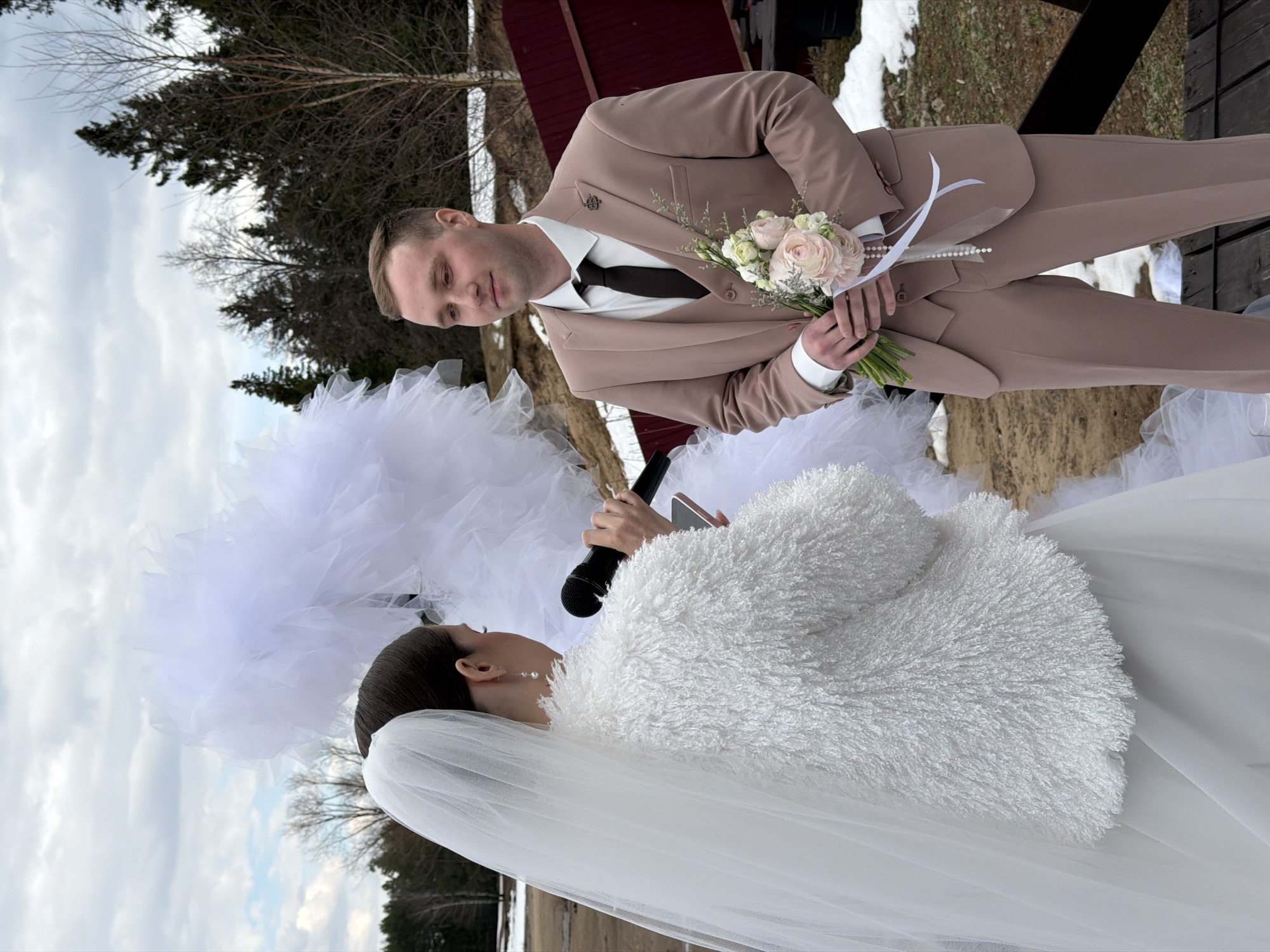 This image captures an outdoor wedding or ceremonial event featuring a bride and groom in Chaikovsky, Russia. The setting suggests late winter or early spring, with patches of snow on the ground, bare deciduous trees, and dark evergreens under a cloudy sky.
The groom, positioned on the right, wears a light brown suit, a white shirt, and a black tie, accented with a lapel pin. He holds a delicate bouquet of light pink and white flowers, adorned with pearls and ribbons, and looks towards the bride with a gentle, attentive expression.
On the left, the bride is seen in a white wedding dress, complemented by a warm white faux fur jacket and a flowing veil over her dark hair. She holds a black microphone, possibly also a smartphone, indicating she might be speaking vows, giving a speech, or performing.
A wooden structure with a red roof is visible in the background. The scene conveys an intimate outdoor celebration despite the cool weather. No text is visible in the image.