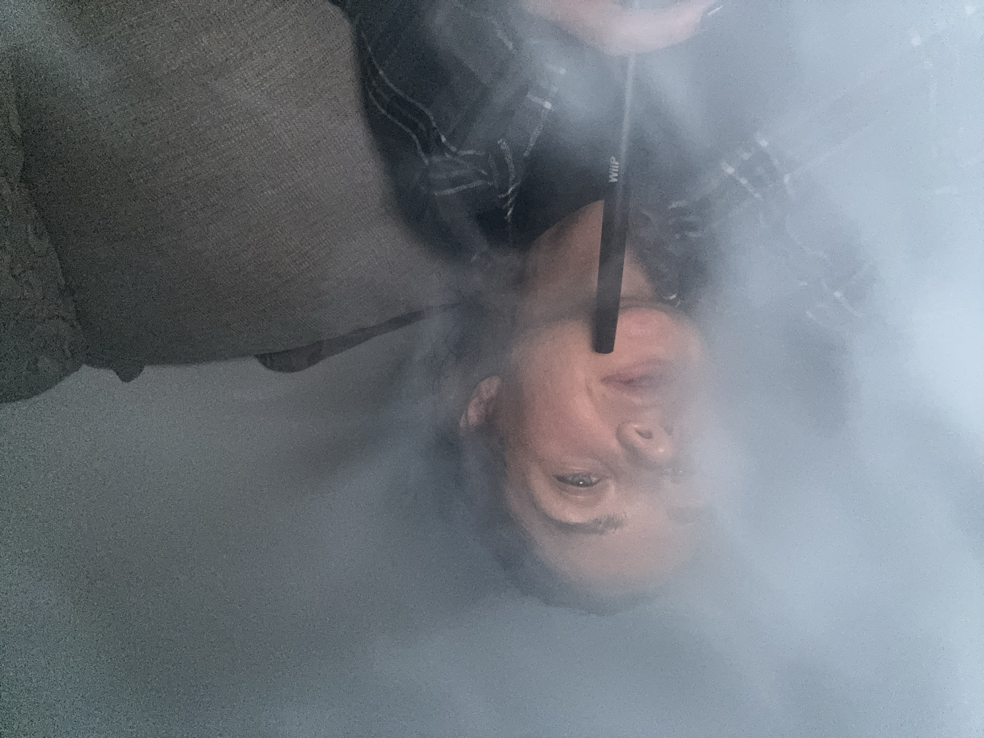 An overhead shot captures a person, likely male, amidst a dense cloud of white vapor or smoke. The individual's head and upper torso are visible, oriented upside down in the frame, with their head positioned towards the bottom of the image. They have fair skin, dark hair, and are looking upwards (towards the top of the frame). One eye is clearly visible, while the other is partially obscured by the smoke.
A slender, dark object is held to their mouth, from which the copious smoke appears to emanate. The word "WHIP" is distinctly visible in white text on this object. The person is wearing a garment with a dark plaid or patterned design, visible on their shoulder. In the background, to the left of the person, is a textured, possibly fabric surface, resembling a couch or cushion, in a muted gray tone. The overall scene is dominated by the smoke, which creates a hazy, soft-focus effect, obscuring clear details of the environment. The activity suggests vaping or smoking a shisha/hookah. The location is an indoor setting, potentially in Budva, Montenegro, though the image itself offers no specific geographical cues beyond the general indoor context.