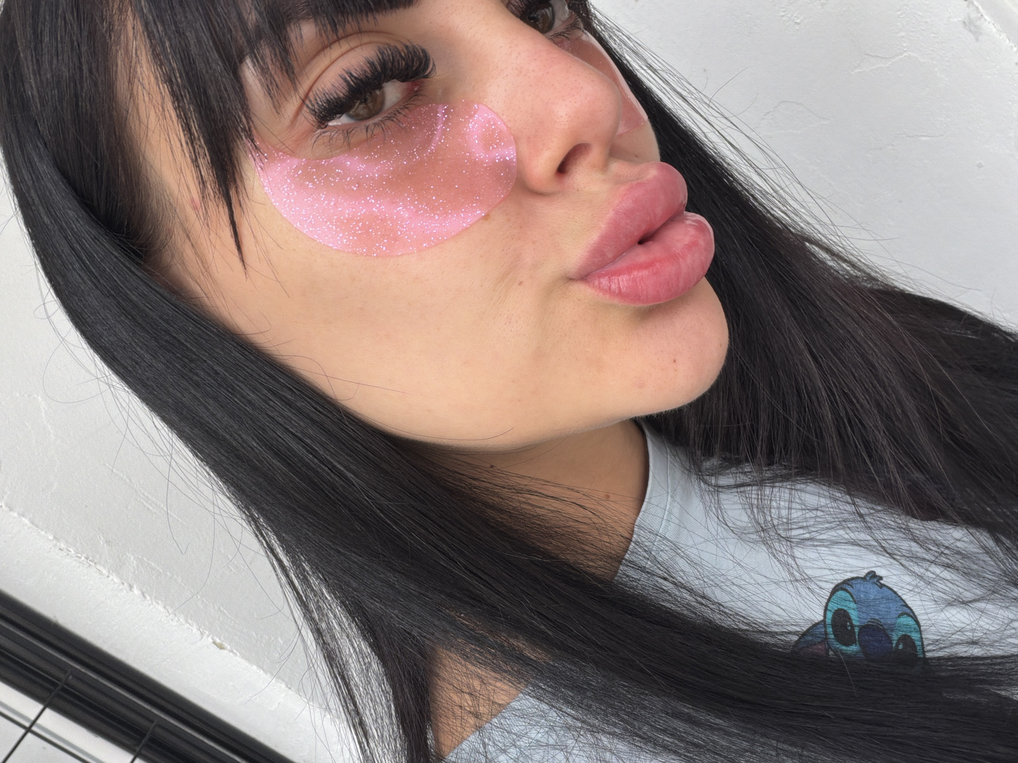 The image shows a close-up portrait of a young woman with long, dark hair and bangs. She is wearing light blue t-shirt with a Stitch character graphic. She has pink under-eye masks on, applied to her cheeks below her eyes, and her lips are slightly parted. The background is a white wall with some texture and a black metal grate in the upper left corner. There is no indication of the location being Plovdiv, Bulgaria.