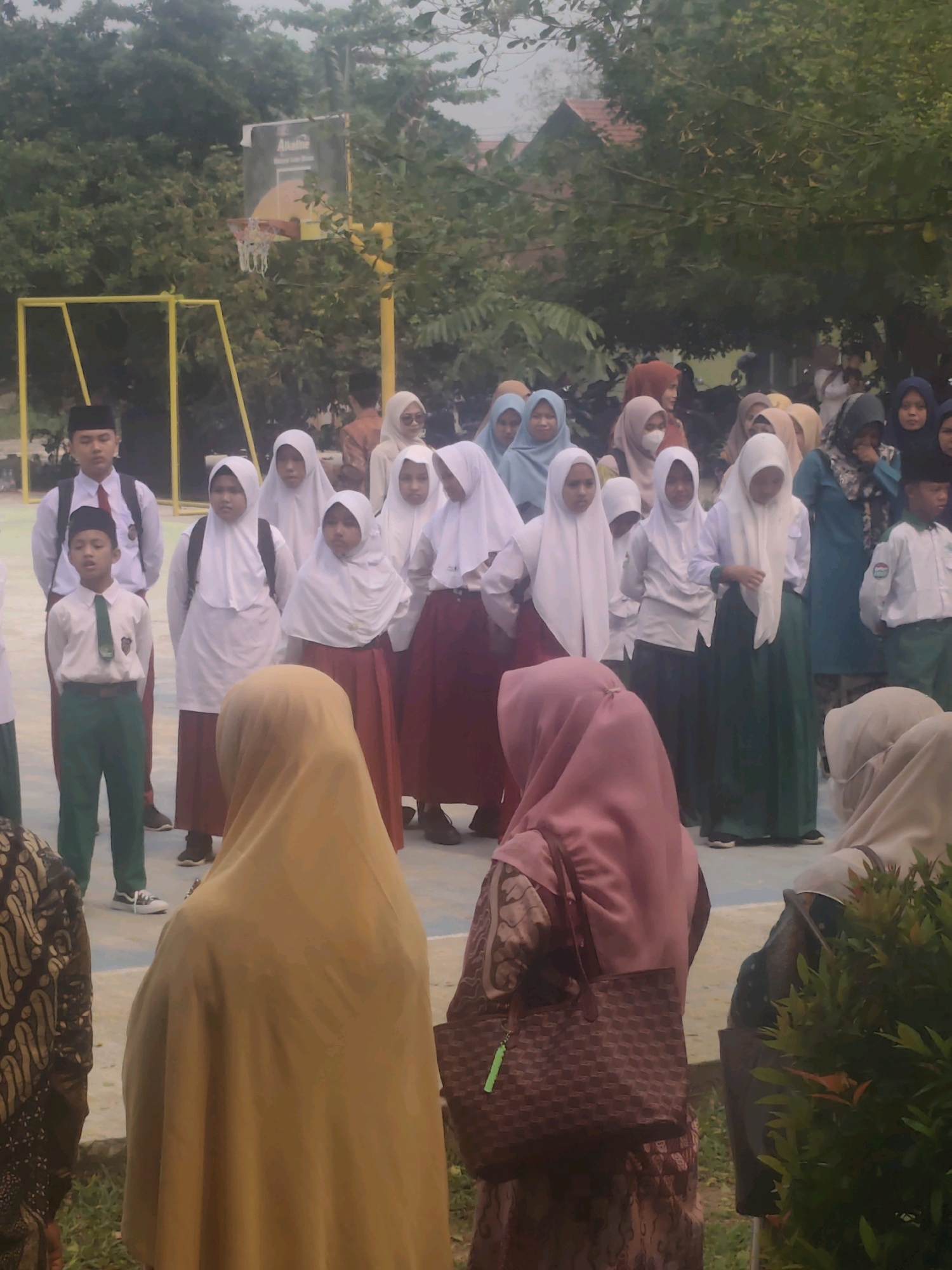 A group of students, predominantly female, are gathered outdoors in what appears to be a schoolyard in Pematang Reba, Indonesia. The students are mostly wearing school uniforms consisting of white shirts and trousers or skirts, often paired with hijabs in various colors, including white, cream, and light blue. Some students are also wearing traditional Indonesian batik clothing. In the background, there is a basketball hoop and pole, and trees and buildings are visible, suggesting an urban or suburban school setting. The overall atmosphere seems to be that of a school assembly or a casual gathering.