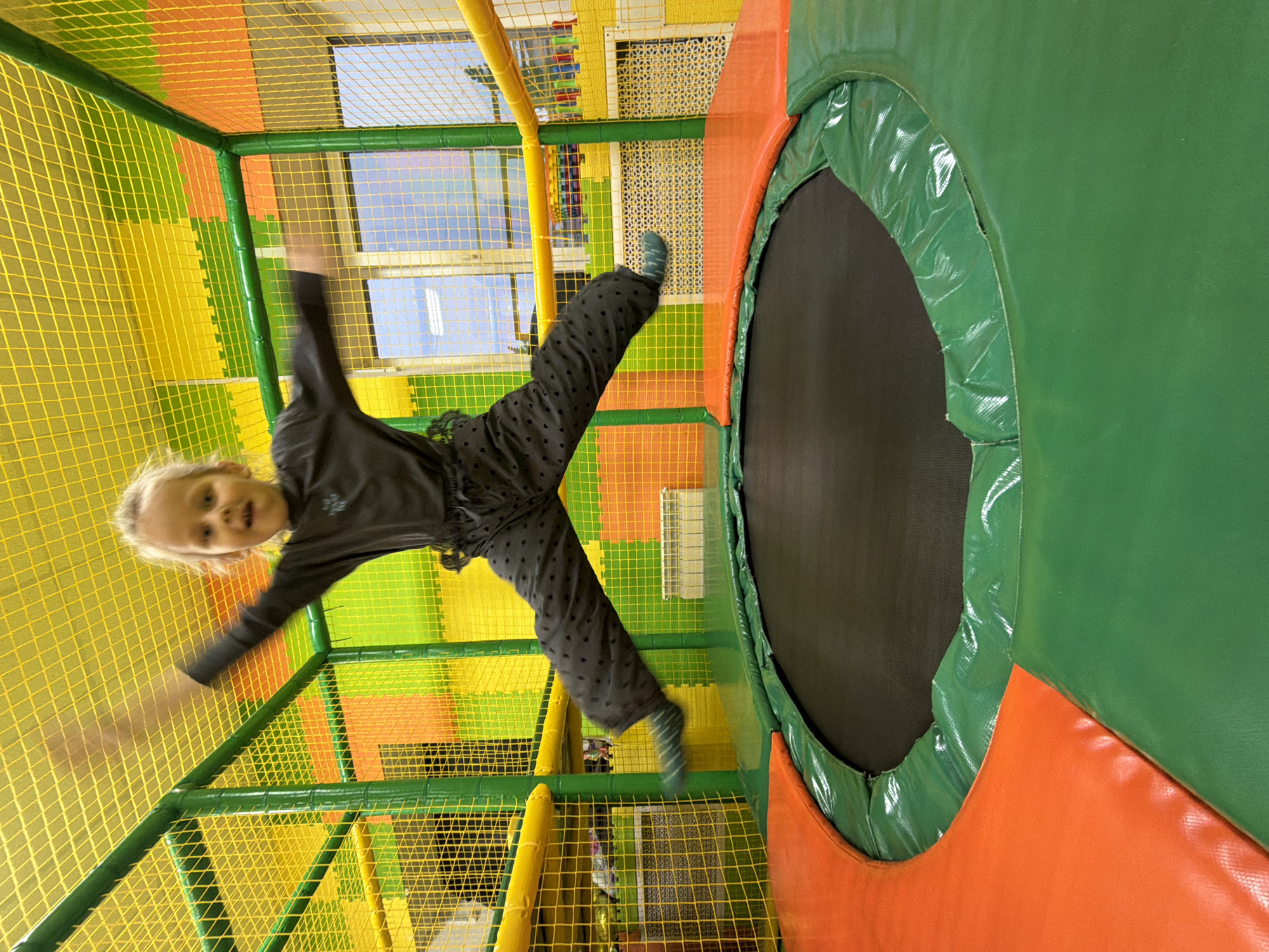 A young child with blonde hair, wearing a dark long-sleeved shirt and patterned dark pants with striped socks, is jumping on a trampoline inside a children's indoor playground. The trampoline has a black jumping surface and is surrounded by green padding with orange and green cushioned walls. The playground structure is constructed with yellow netting and green frames. The environment includes colorful padded flooring, and a window is visible in the background. The scene appears to be an indoor entertainment area, possibly located in Kamyshin, Russia. The child's arms are outstretched, suggesting active movement during a jump.