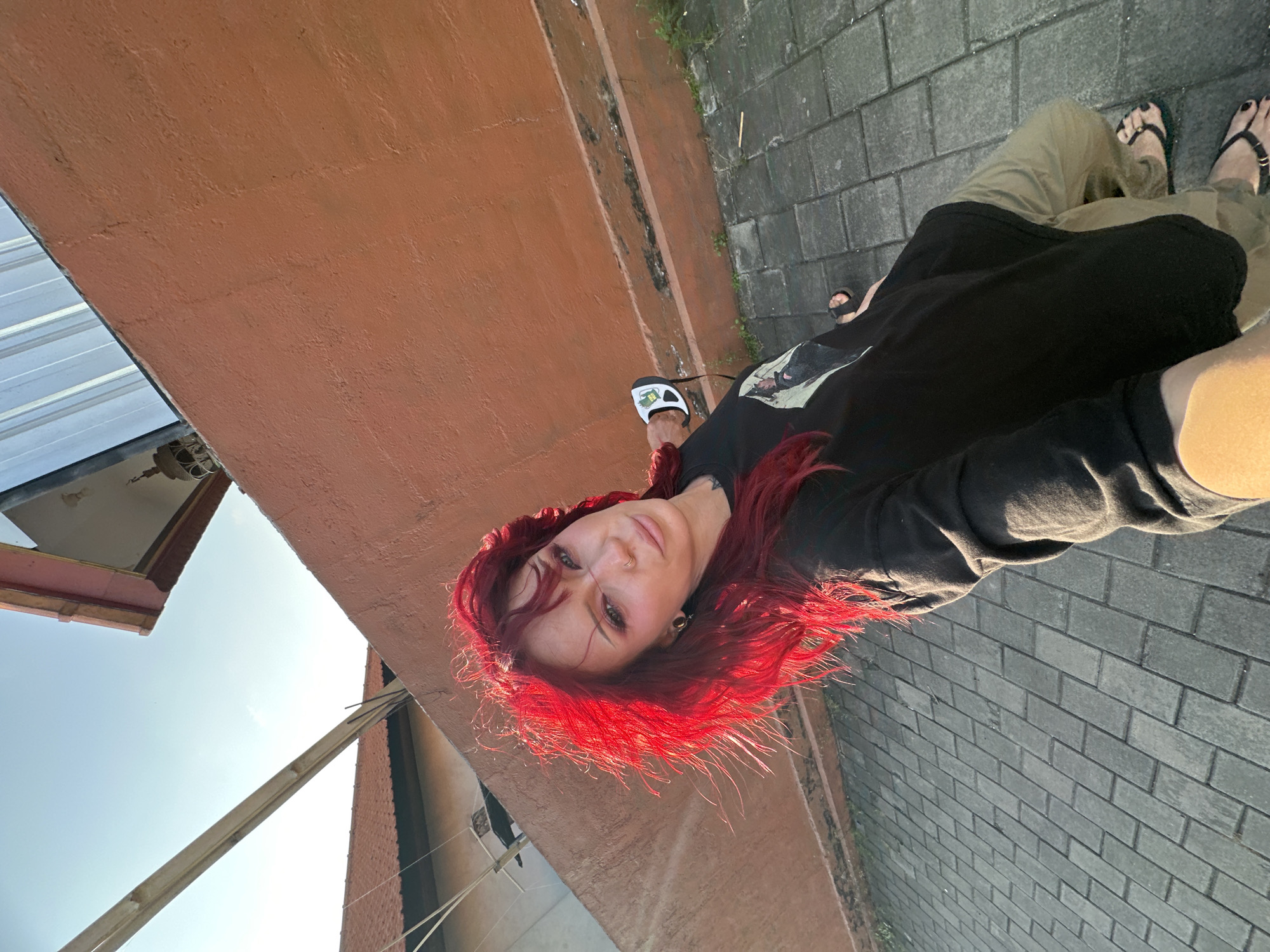 A woman with bright red hair is standing on a paved walkway outside, holding a dog leash in her right hand. She is wearing a black long-sleeved shirt and tan pants. She is looking towards the camera with a neutral expression. The background consists of a textured orange wall and a blue sky. The scene appears to be located in North Kuta, Indonesia.