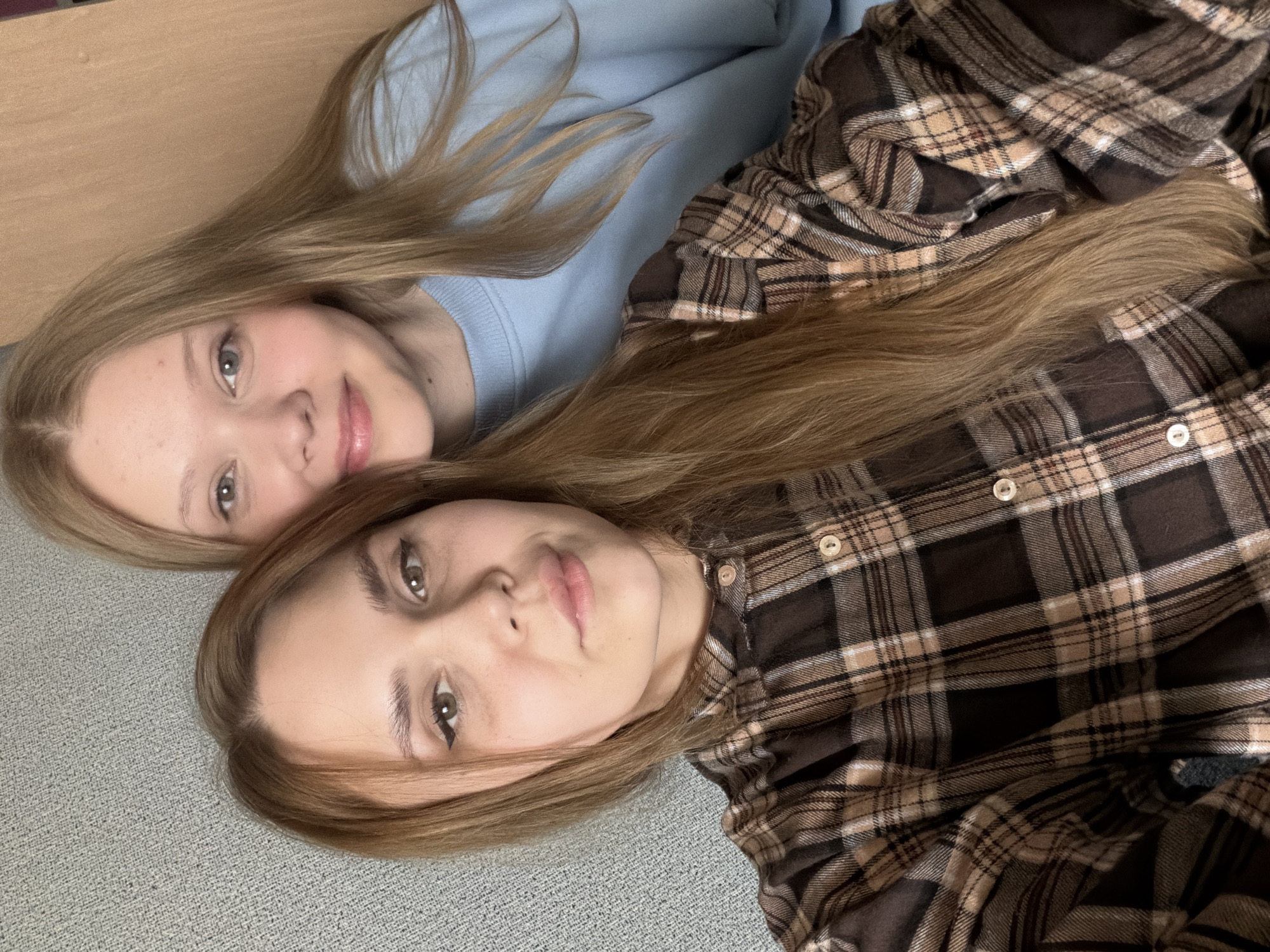 Two young women are lying on a light gray carpet. The woman on the left is wearing a light blue sweatshirt, while the woman on the right is wearing a brown and tan plaid shirt. The location is likely indoors, but there is nothing to indicate that it is specifically in the city of Chaikovsky, Russia.