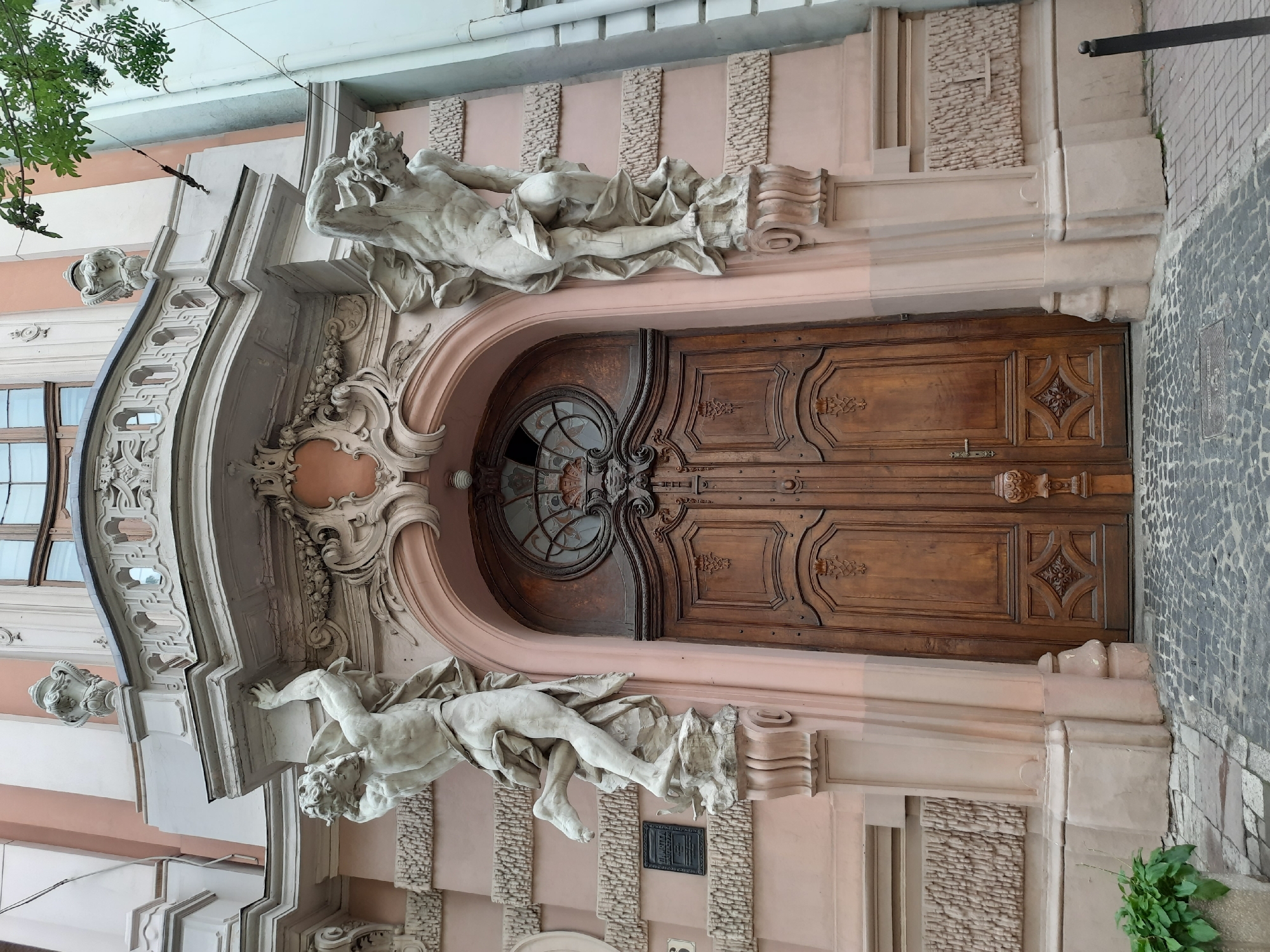 The image shows the ornate wooden double doors of a building with elaborate sculptures flanking the entrance. The sculptures depict male figures in dynamic poses, seemingly supporting the balcony above. The building's facade is painted a pale pink with white architectural details. Above the doors is a decorative oval window with intricate metalwork. The ground in front of the building is paved with cobblestones. The scene is likely located in Lviv, Ukraine, given the architectural style which is characteristic of the city. The overall impression is one of historical grandeur and architectural artistry.
