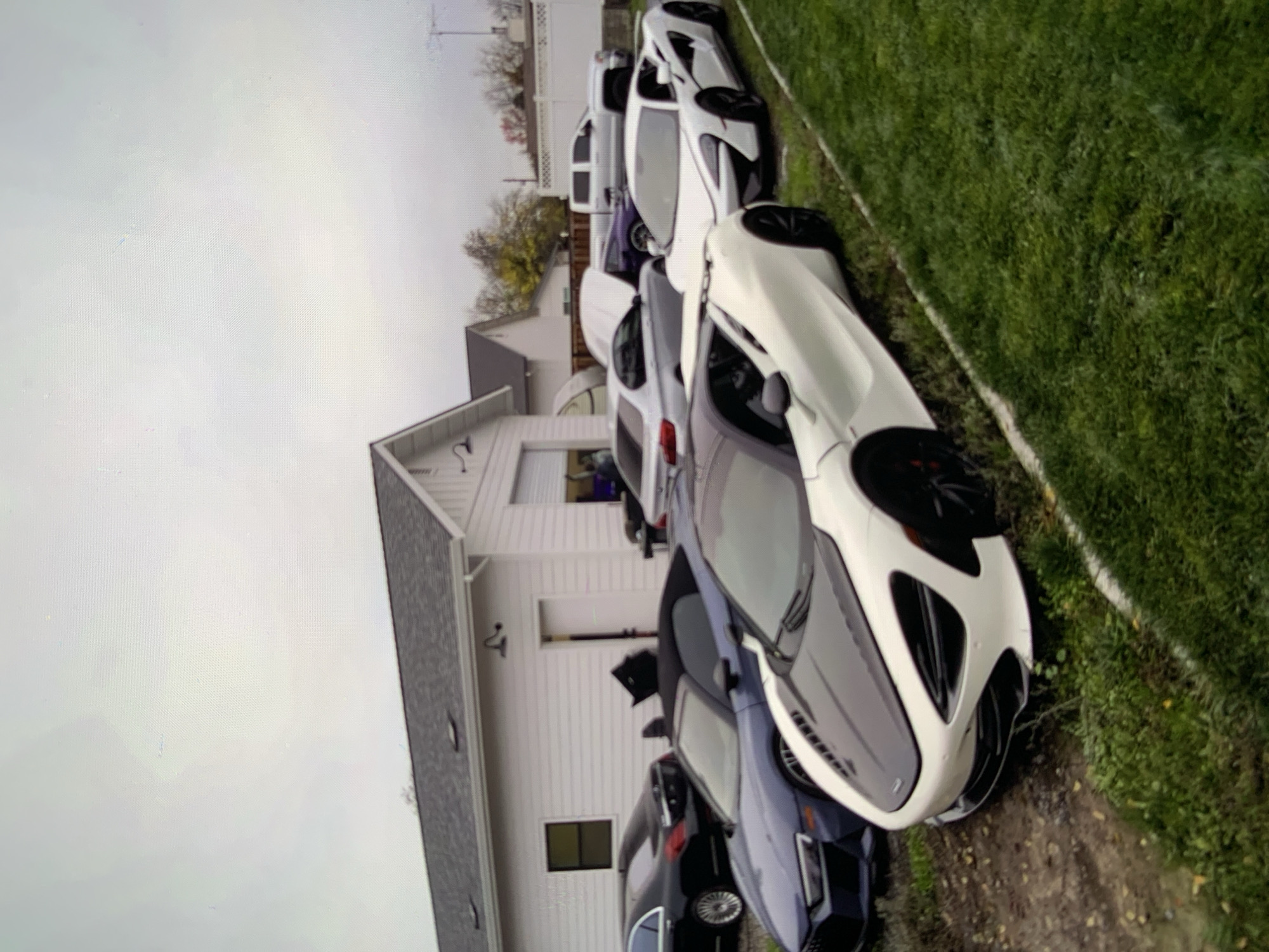 In an outdoor setting, several luxury vehicles are parked next to a white building with a dark gray roof. The building has a large garage door and a smaller side door. In the foreground, a white McLaren 720S is parked on the right, with its front end angled towards the viewer. To the left of the McLaren, a dark blue convertible sports car, possibly an Audi R8, is parked. Further back, a black sedan, likely a Mercedes-Benz S-Class, is visible. Behind these cars, a silver coupe with its hood open is parked next to a dark purple vehicle. To the right of the building, a silver pickup truck is parked. The ground is a mix of dirt and grass. The sky is overcast.