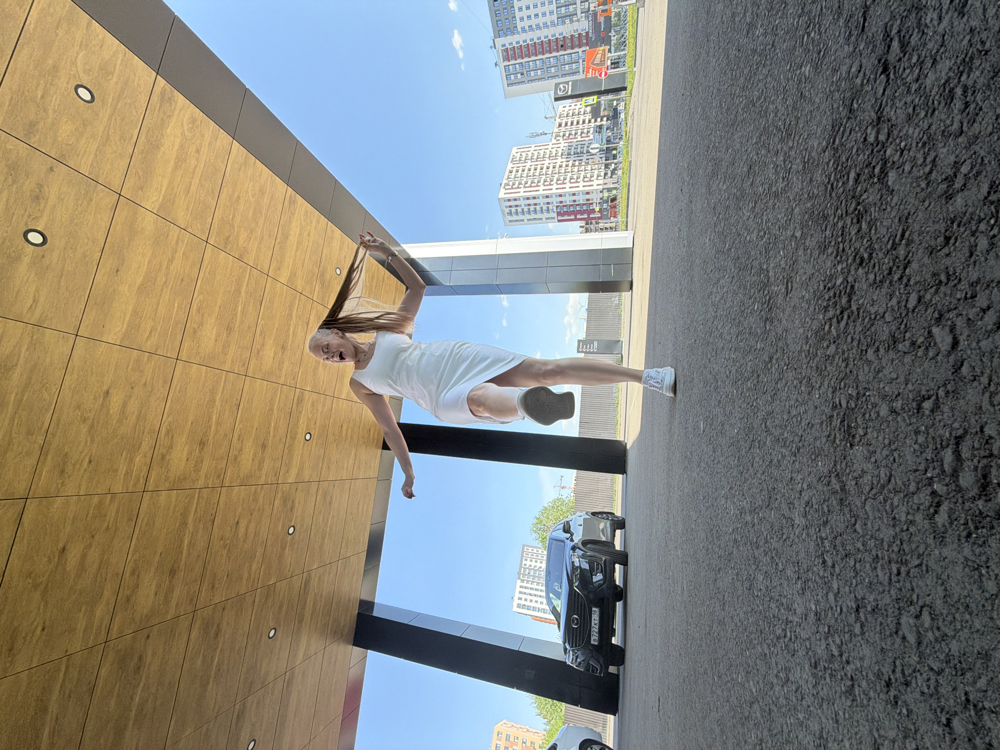 A medium shot captures a fair-skinned woman in mid-air, clad in a white dress and sneakers, amidst an urban environment in Izhevsk, Russia. She appears to be playfully pulling her long, light-brown hair upwards, her mouth open in an expression of enjoyment or surprise.
The background reveals a modern cityscape, with several high-rise buildings under a clear blue sky. A black SUV is parked nearby. The ground is asphalt. The woman is positioned near a building with a distinctive wood-panel facade.