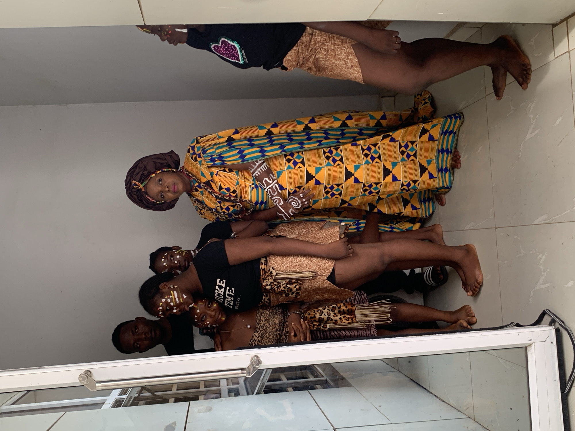 A group of six people are reflected in a glass window or mirror. The people are dressed in a mix of modern and traditional African clothing. One woman wears a brown head wrap and a yellow and blue patterned robe. Other people are in black T-shirts, leopard print accessories, and brown shorts. Some have white paint on their faces. The background appears to be a room with white walls and a tiled floor. Based on the prompt, the location is in Akwanga, Nigeria, though specific landmarks or details to confirm this are not visible in the reflection.