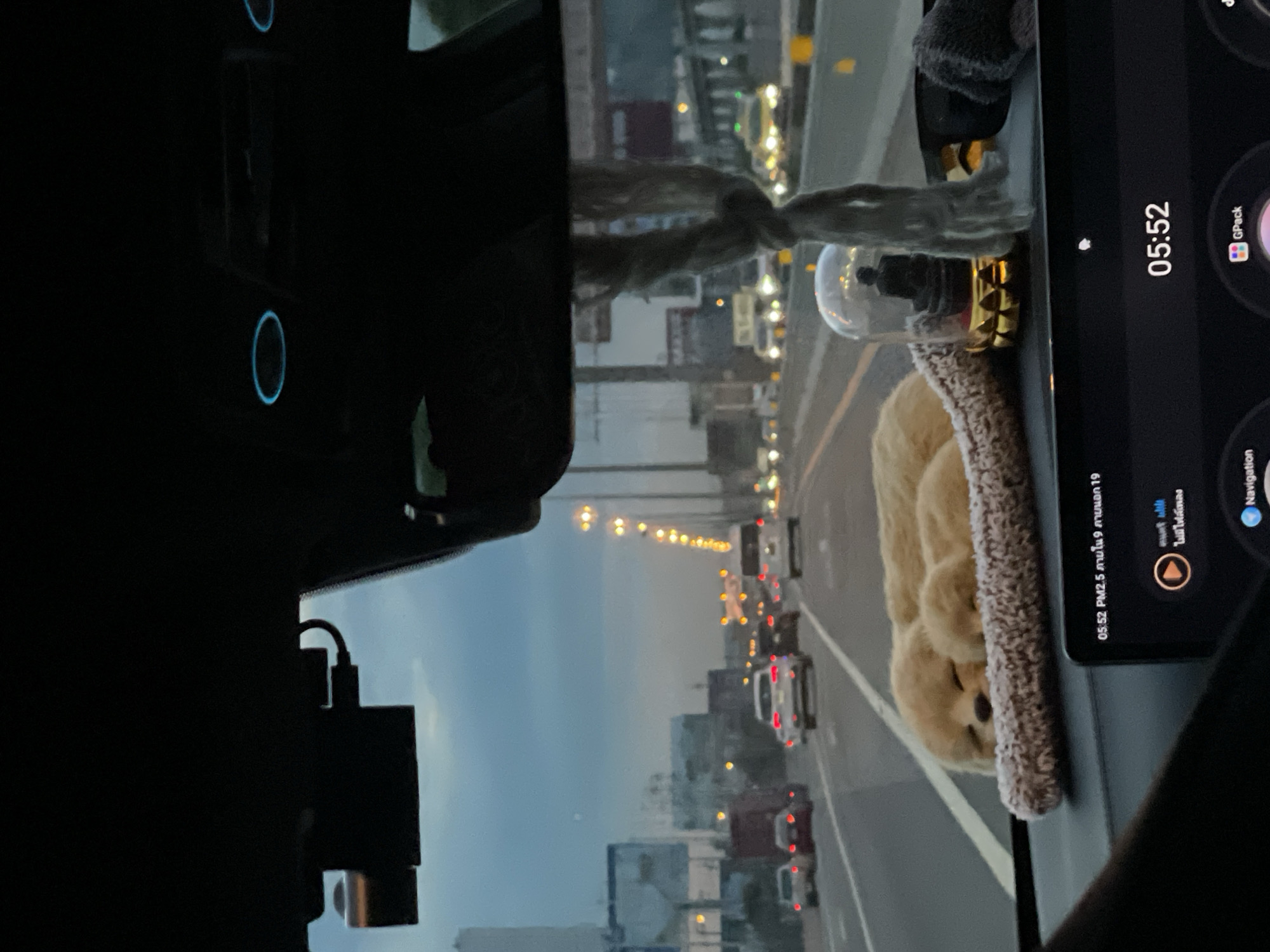 The image shows a view from inside a car driving in Bangkok, Thailand at 05:52 AM. The car is on a highway with heavy traffic, as seen through the windshield. On the dashboard, there is a teddy bear and a snow globe placed on top of a tablet displaying Thai text and icons related to navigation and air quality ("PM2.5"). The sky is overcast. A dashcam is affixed to the windshield.