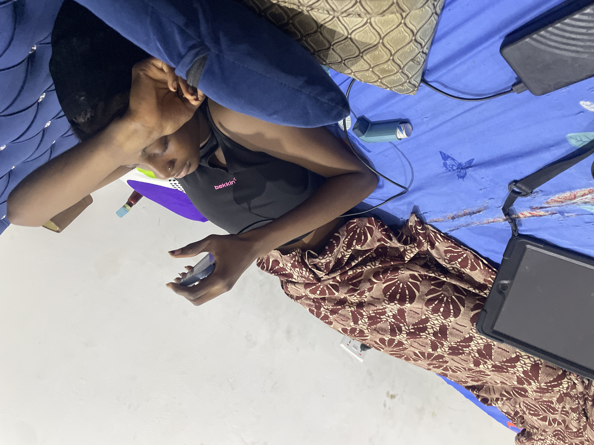 The image shows a young Black woman lying on a bed in a room that appears to be in Abuja, Nigeria. She is looking at a mobile device in her hands. Her right arm is resting on her head, and her left arm is bent with her hand supporting the device. She is wearing a black tank top and a patterned wrap skirt.
The bed is covered with a blue sheet that has some subtle printed designs. To the left of the woman, on the wall, is a white electrical outlet. Beside the woman, on the bed, is a teal blue inhaler and a black tablet with a ruggedized case. A black cable runs from near the inhaler towards the bottom right of the frame where it connects to a black power bank. A portion of a dark blue upholstered headboard is visible at the top of the image. The wall behind the bed is white and shows some discoloration and marks. The overall lighting suggests the image was taken indoors, possibly at night or in a dimly lit room.