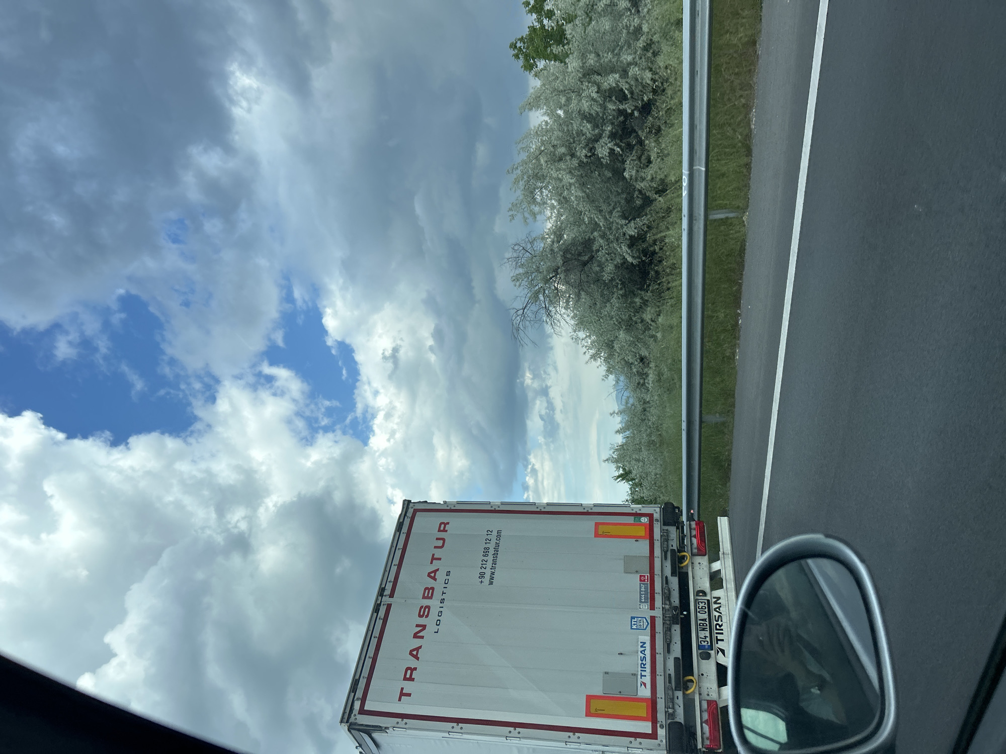The image depicts a view from inside a vehicle traveling on a highway or major road. A large white truck with red lettering reading "TRANSBATUR LOGISTICS" is directly in front of the vehicle. Contact information for the company is also visible on the back of the truck, including a phone number and website. The truck's license plate reads "34 NBA 063". To the right of the truck, there is a guardrail and a stretch of trees or bushes with light-colored foliage. The sky above is partially cloudy, with patches of blue sky visible. Part of the car's side mirror is visible in the foreground of the image.
The location is not Malo Konare, Bulgaria based on license plate information. The license plate is a Turkish license plate.