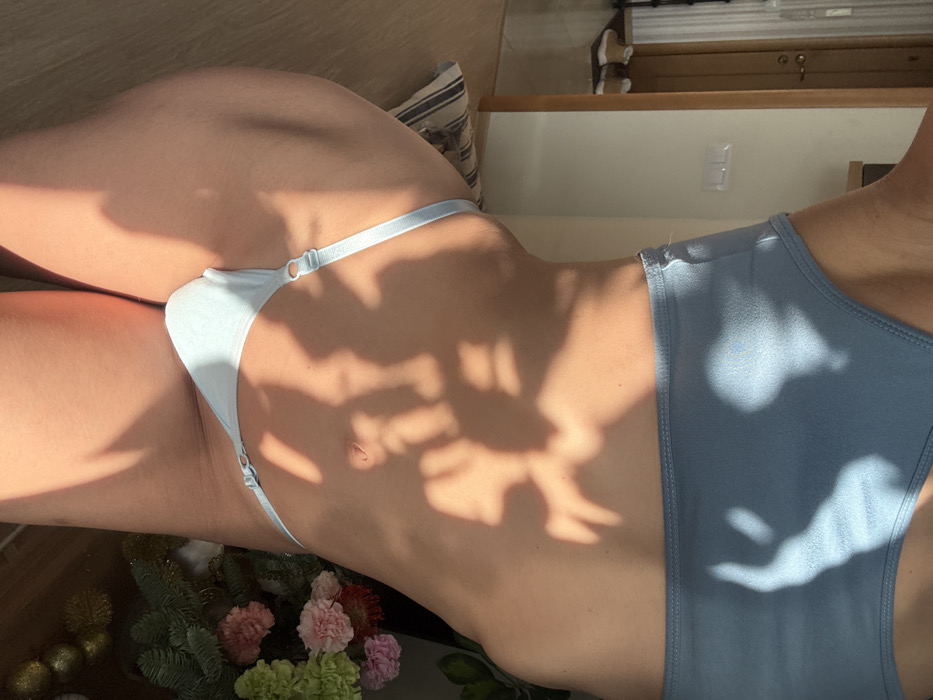 The image presents a close-up, mid-torso view of an individual, likely female, from the lower chest to the upper thighs, within an indoor domestic setting. The person wears a light blue athletic crop top or bra and matching light blue thong-style underwear featuring adjustable side straps. Direct sunlight illuminates the scene, casting distinct, dendritic shadows, consistent with foliage, across the person's skin and portions of the background.
To the left of the subject, a light-colored wooden door frame is visible, opening to a hallway or adjacent room with a light wood floor. On this floor, two pairs of light-toned casual shoes are placed. A white wall-mounted triple light switch is visible on the wall adjacent to the door frame. A striped cushion or pillow is partially visible on the floor near the subject's left hip. To the right of the subject, a floral arrangement containing pink and green flowers is situated. Below the flowers, several golden, spherical ornaments are present on a surface or the floor. The environment is an interior space with no external features or landmarks identifiable to confirm a specific location within Saint Petersburg, Russia. No explicit actions or interactions are depicted beyond the subject posing for the photograph.