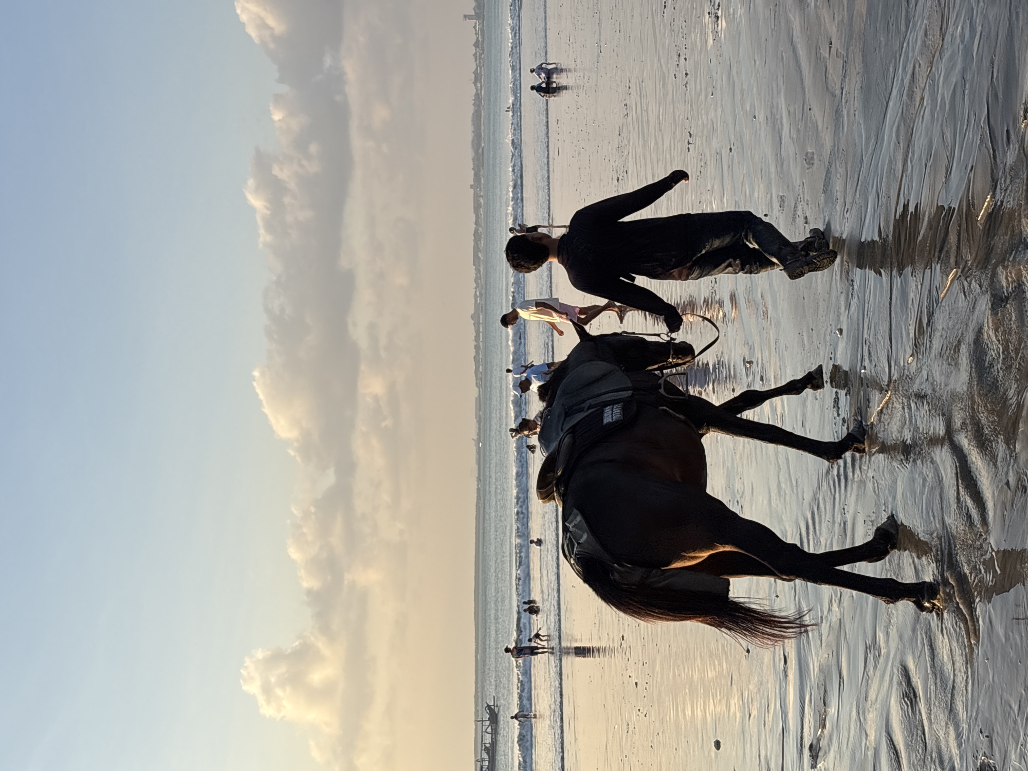 The image captures a beach scene in Kuta Selatan, Indonesia, under a warm, diffused late afternoon or early evening light. In the immediate foreground, a man, viewed from behind and wearing dark long-sleeved attire and boots, walks towards the right side of the frame, leading a dark-colored horse by its reins. The horse is fitted with a saddle and walks alongside the man, its hooves creating ripples in the shallow, reflective water at the ocean's edge.
The background features numerous people dispersed across the wet sand and in the ocean. Several individuals are seen wading, standing, or playing in the gentle waves, while others appear to be relaxing closer to the water's edge. On the far left, a traditional wooden boat is partially visible in the ocean. The sky transitions from a light blue overhead to a warm, golden-orange hue near the horizon, populated by scattered, illuminated cumulus clouds. A distant coastline with a low-lying urban landscape and a prominent tower structure is discernible on the horizon line. The wet sand reflects the sky and the figures, emphasizing the golden hour lighting.