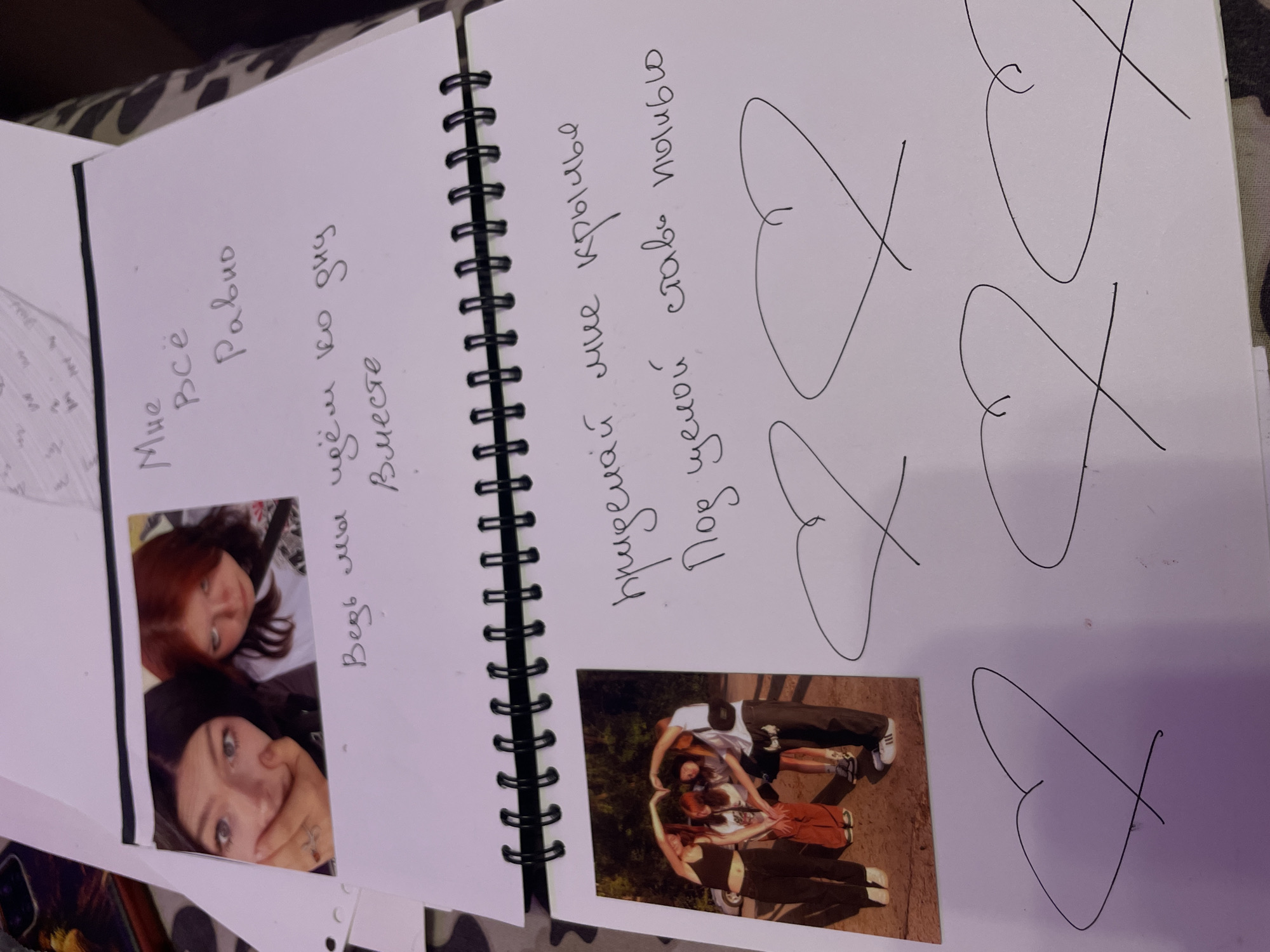 This image depicts a page in a spiral-bound notebook. On the upper half of the page, a photograph features two young women. One woman on the left has dark hair and is covering her mouth with her hand, revealing a ring on her finger. The woman on the right has reddish-brown hair and is looking towards the camera with a neutral expression. Text in Russian is handwritten next to the photograph. The text reads, "Мне всё равно" (Mne vsyo ravno), which translates to "I don't care." Below this, more text reads, "Ведь мы идём ко дню вместе" (Ved' my idyom ko dnyu vmeste), meaning "After all, we are walking towards the day together."
The lower half of the page contains another photograph, this one featuring four individuals standing outdoors, forming a heart shape with their arms. Below this photograph, more Russian text is written: "придемаи мне крылья" (pridemai mne kryl'ya), translating to "give me wings," and "под ценой ставь пылью" (pod tsenoy stav' pyl'yu), which translates to "under the price put dust." Scattered across the bottom of the page are several hand-drawn heart shapes. The notebook appears to be resting on a patterned surface, possibly a blanket or fabric. The location is not specified as Vaganovo, Russia.