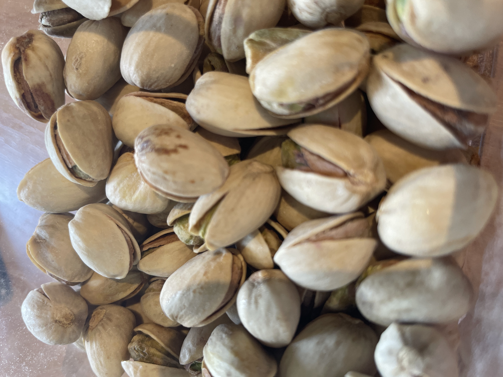 A close-up, overhead shot shows a pile of shelled pistachios. The pistachios are light tan in color with some having a reddish-brown hue. The shells are partially opened, revealing the green nuts inside. The nuts are densely packed together, filling the frame of the image. The lighting is bright and even, suggesting an indoor setting. There are no people or text visible in the image. The focus is entirely on the texture and arrangement of the pistachios. The scene appears to be a simple still life of food.