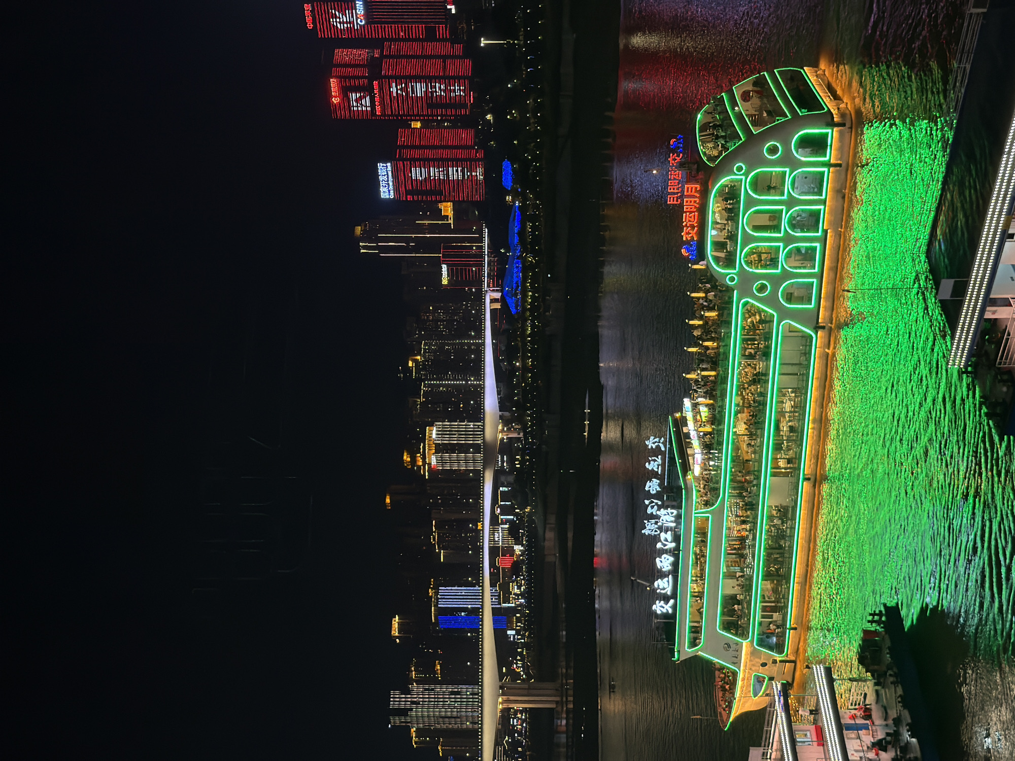 This is a nighttime, vertical, medium-shot photograph of a riverfront cityscape in the Yuzhong District of China. The scene is illuminated by the bright lights of buildings and a brightly lit tour boat.
The main subjects are the city skyline and a large tour boat. The skyline features numerous tall buildings with various lights, including red, white, and blue illumination, creating a vibrant urban backdrop. Some buildings have prominent signage, with text such as "CASINO" and "China Development Bank" clearly visible.
The tour boat, the "Jiaoyun Liangjiang You," is a dominant feature on the right side of the image. It is outlined and decorated with bright green neon lights, making it stand out against the dark water. The boat appears to be on a river, and its green lighting casts a vibrant glow on the water's surface. The boat's interior is visible, showing people inside, suggesting it is in operation and carrying passengers.
The time of day is clearly night, as indicated by the darkness of the sky and the reliance on artificial lighting for visibility. The water is calm, reflecting the city lights and the boat's illumination. There are no clear indicators of weather, but the scene suggests a clear night.
Visible text includes "交运两江游," "民即交、" and "交运明月" on the boat, and "中国平意," "CASINO," and "国家开发银行" (China Development Bank) on the buildings. The overall mood is one of vibrant city life and nighttime entertainment.