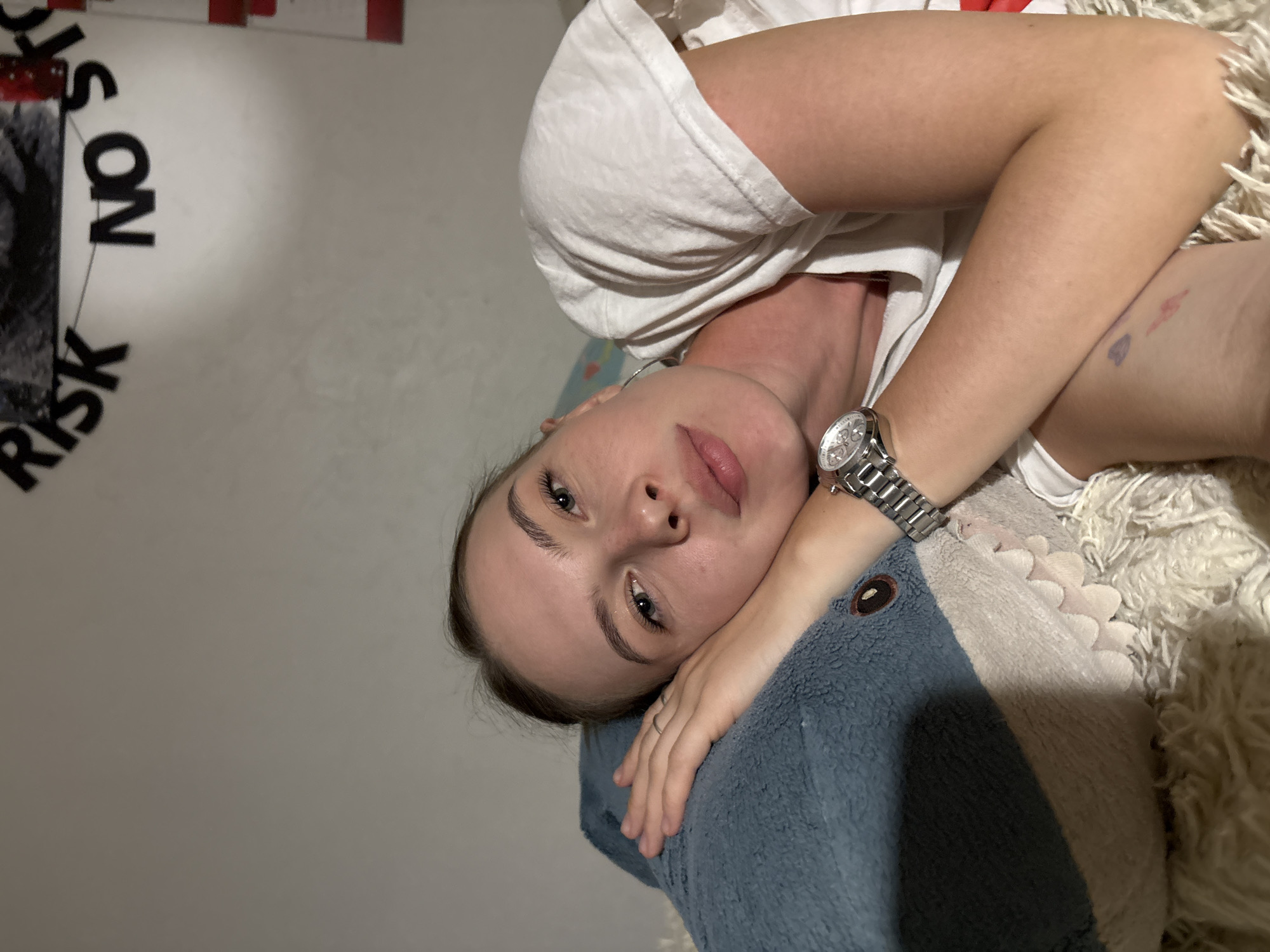 The image shows a young woman lying on her side on a plush, white rug, resting her head on a large, blue shark stuffed animal. She is wearing a white t-shirt and a watch on her left wrist. Her right arm is bent, with her hand resting on the shark. The background is a plain wall, with a banner displaying the words "RISK NO" visible at the top. The scene appears to be indoors, likely in a bedroom, and there is no indication of the city of Perm, Russia.