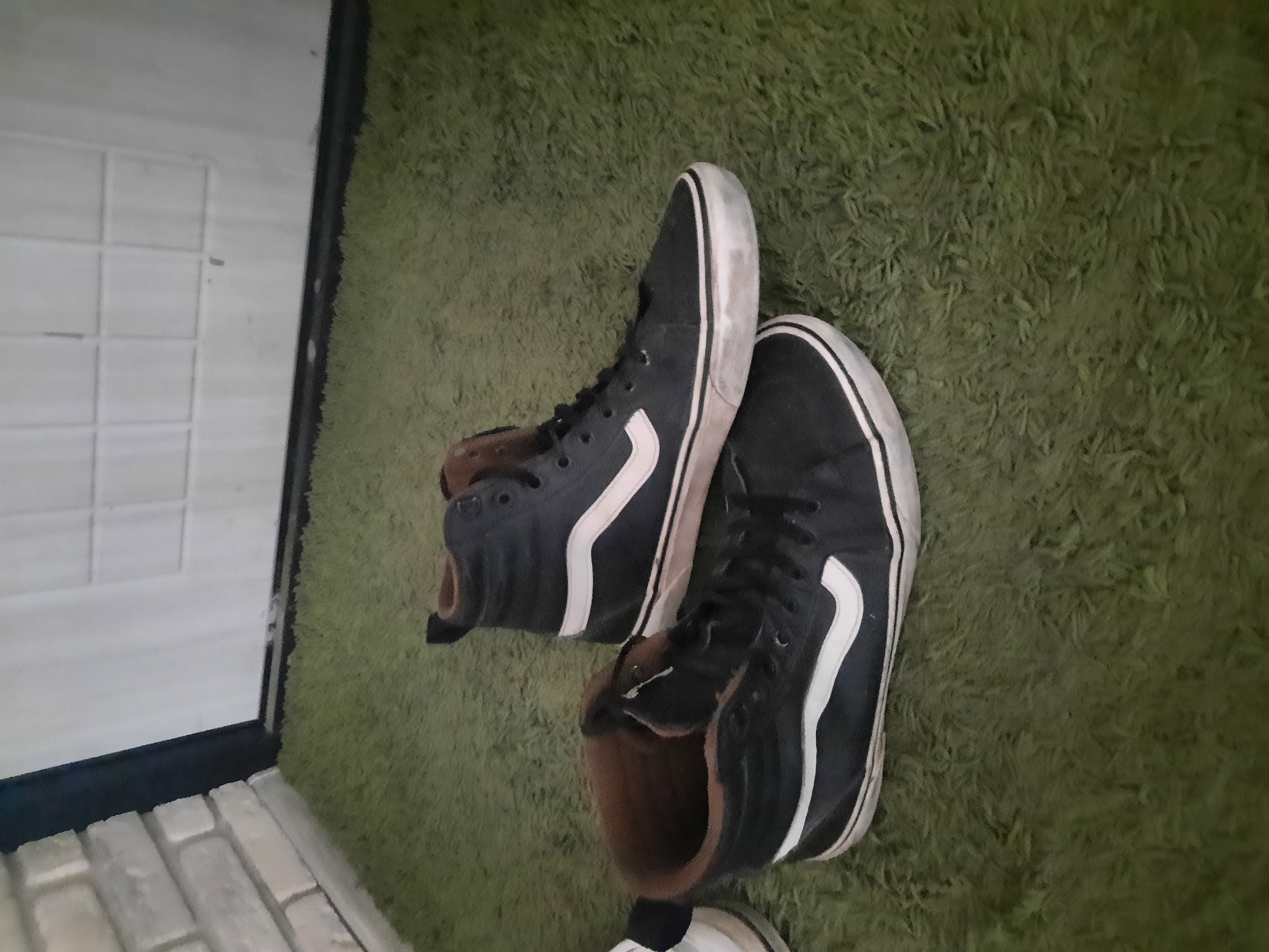 In an indoor environment with a green shag carpet, a pair of dark blue high-top Vans sneakers with white accents are visible. One shoe is positioned in front of the other, with the rear shoe slightly elevated. The sneakers appear worn. To the left of the sneakers, a portion of a white brick wall can be seen, and to the right, a white door with paneling is present. The lighting in the scene is dim.