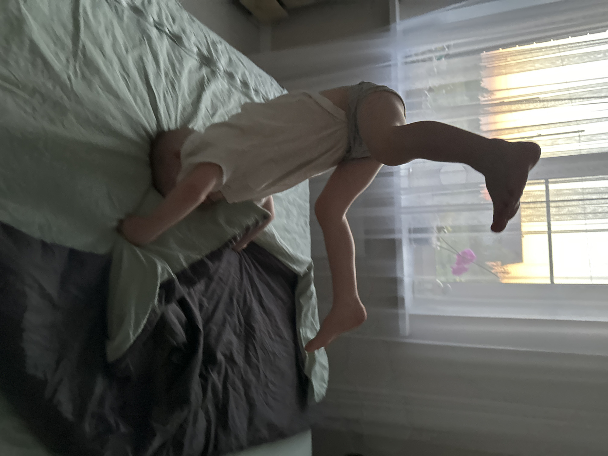 A child wearing a white t-shirt and underwear is bent over a bed. The child's head and one arm are on the bed, while the other arm and both legs are extended off the bed. A window with sheer curtains is in the background, with blinds visible behind the curtains. A flower can be seen outside the window. The location is indoors and the place name is Прикамский, Russia.