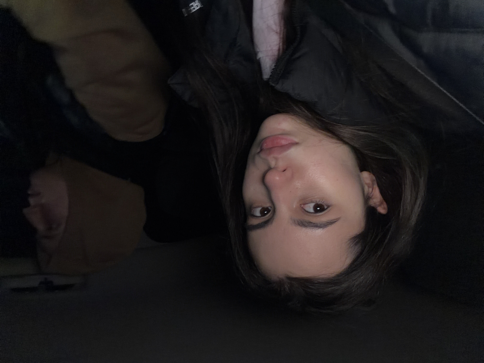 This media file is a close-up portrait, rotated 90 degrees counter-clockwise. The primary subject is a young woman, likely in her late teens or early twenties, with long, straight dark brown hair. She is looking directly at the camera with dark brown eyes and a neutral expression, her lips slightly parted. Her skin appears smooth and clear.
She is wearing a black puffer jacket, visible across the upper right portion of the frame (when oriented correctly), beneath which a hint of a light pink or white top can be seen. There is faint, unidentifiable white text on the black jacket, possibly a brand logo.
The background is predominantly dark and out of focus. In the upper left quadrant of the rotated image, a blurry, indistinct figure or reflection of another person is visible, wearing a light brown or tan garment, possibly a hoodie, largely obscured by shadows. The rest of the background is a dark, uniform space, suggesting an indoor setting with low light or a nighttime outdoor environment.
The lighting is soft and appears to be coming from the front, illuminating the woman's face and creating subtle shadows. The overall mood is calm and somewhat intimate. The activity is a posed photograph. No specific location cues are visible within the image, but the provided context indicates the location is Chaikovsky, Russia.