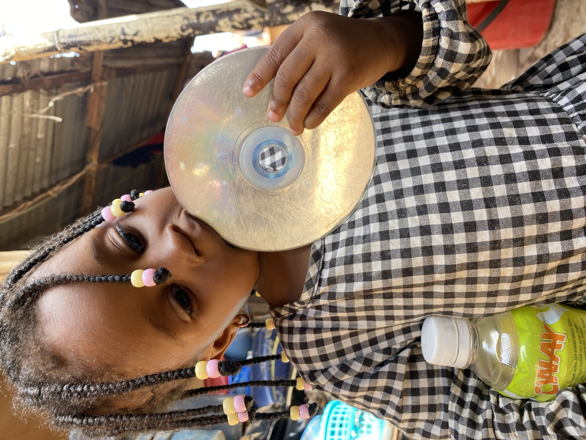 In Bukuru, Nigeria, a young Black girl with braided hair adorned with pink and yellow beads is shown holding a CD close to her face. She is wearing a black and white checkered shirt. Her hair is braided into multiple strands, with some braids framing her face and others falling around her shoulders. She has dark eyes and appears to be looking slightly upwards. A plastic bottle with a yellow and green label is visible in the lower right of the frame, partially obscured by her shirt. The background suggests an outdoor or semi-outdoor setting with wooden structures and corrugated metal roofing, possibly indicating a humble dwelling or workshop. The lighting suggests it is daytime. The label on the bottle contains the text "CALAY". The overall impression is a candid moment of a child playing with an object in a simple, rustic environment.