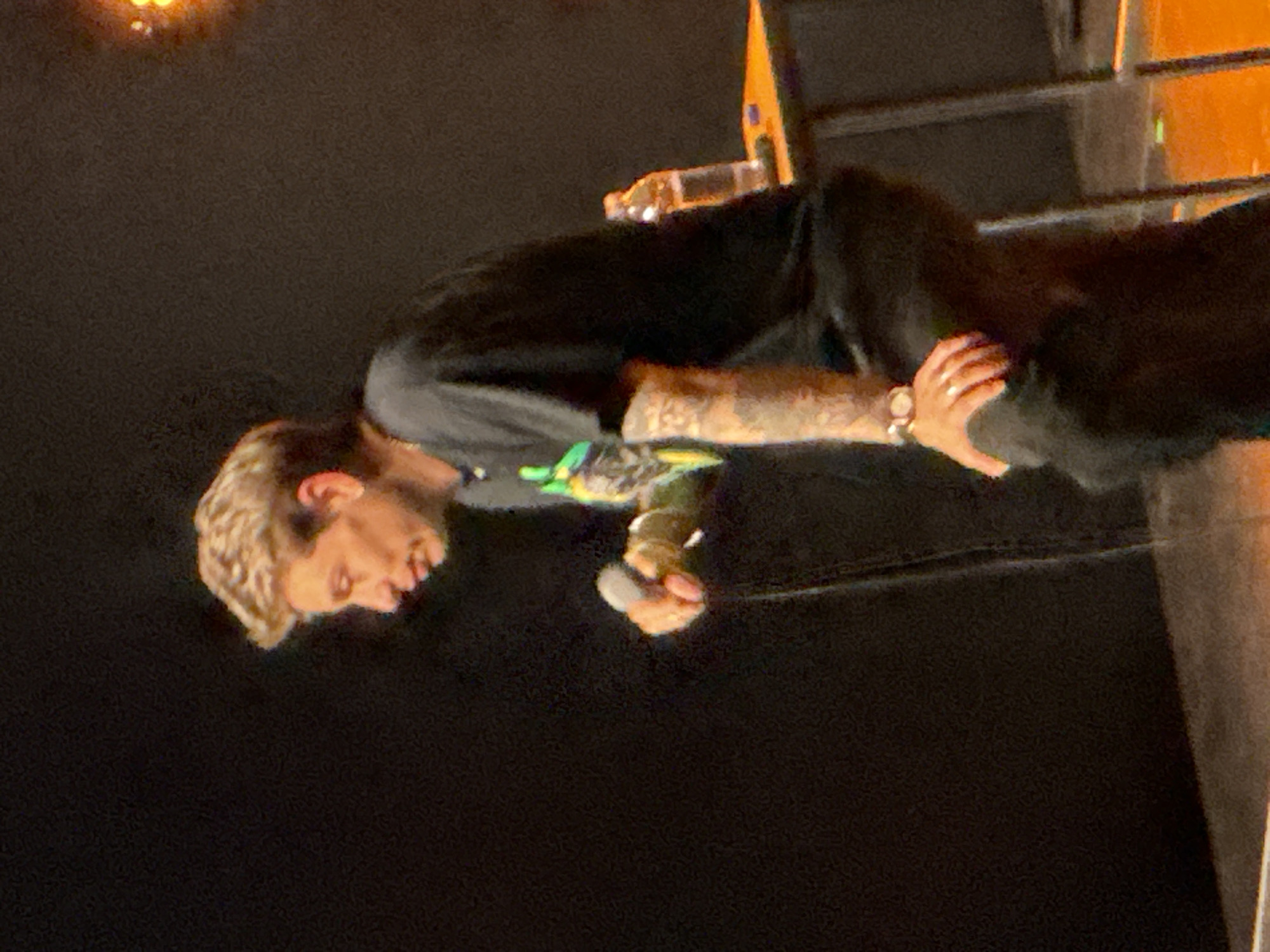 This is a low-light, indoor photograph capturing a male performer on what appears to be a stage. The subject, viewed from the waist up, has light-colored hair and is dressed in a dark long-sleeved top over a lighter shirt with green and yellow graphic elements. Visible tattoos adorn his forearms, and he wears a watch on his left wrist. He is holding a microphone in his right hand, with his head tilted downwards and his mouth slightly open, suggesting he is singing or speaking.
The setting is dark, indicative of an evening or night event, likely a concert, club, or performance venue. The background features indistinct dark areas with glimmers of orange and yellow light, possibly from stage lighting or props. A clear bottle is visible on a raised surface behind the performer. The image conveys an atmosphere of a live musical or spoken word performance. No visible text is present in the image. The provided location context is Vnukovo, Russia.