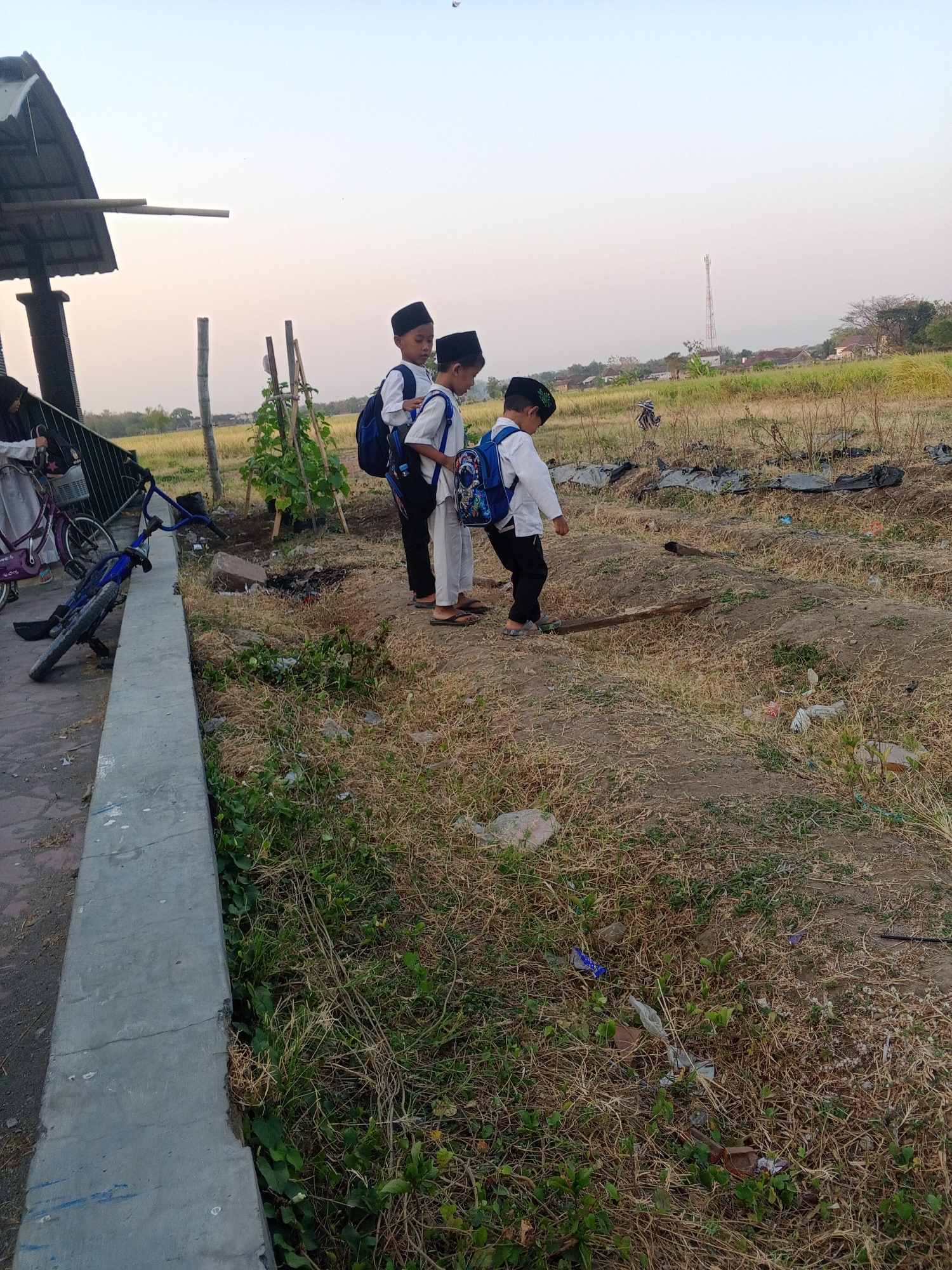Three young boys, dressed in white shirts, black pants, and traditional peci caps, are standing in an outdoor area. They are all wearing backpacks. The boy on the left is facing away from the camera, the middle boy is looking down, and the boy on the right is also looking down and slightly to the side. In the background, there are fields, trees, and a distant cell tower. To the left of the boys, there is a covered structure with a metal roof and wooden supports, and a fence. A blue bicycle is lying on its side near the fence. The ground is covered in dry grass and some green vegetation, with scattered debris. The scene appears to be in a rural or semi-rural area, likely in Lembah, Indonesia, given the traditional attire and the environment.