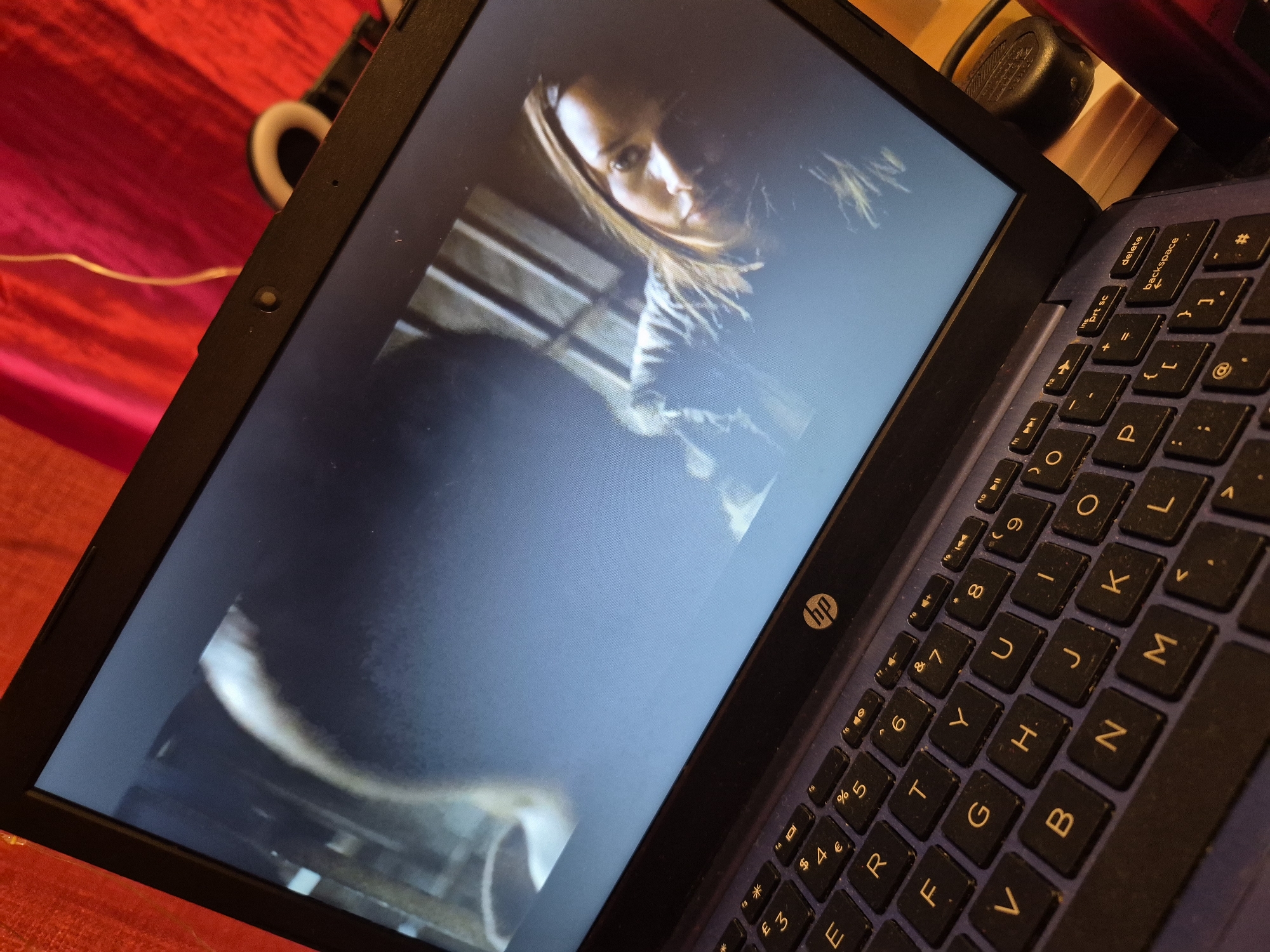 A low-angle, eye-level shot shows a dark blue HP laptop open and displaying a scene from a movie. The laptop is placed on a surface that appears to be covered by a red, textured fabric, possibly a blanket or curtain, suggesting an indoor setting. A thin, yellow cord, possibly a charger or earphone cable, is visible in the upper right corner of the image, trailing across the red fabric. The laptop's screen shows two figures: a person's head in profile on the left, out of focus, and a woman's face in the center-right, also partially obscured by shadow. The woman has dark hair and is looking towards the viewer, her gaze intense. Behind her, vertical bars suggest a cage or a similar structure. The keyboard of the laptop is visible in the foreground, with its black keys and silver lettering. The HP logo is present on the laptop's bezel. The scene appears to be a still from a drama or thriller film. There is no indication of the city of Sandwell, United Kingdom.