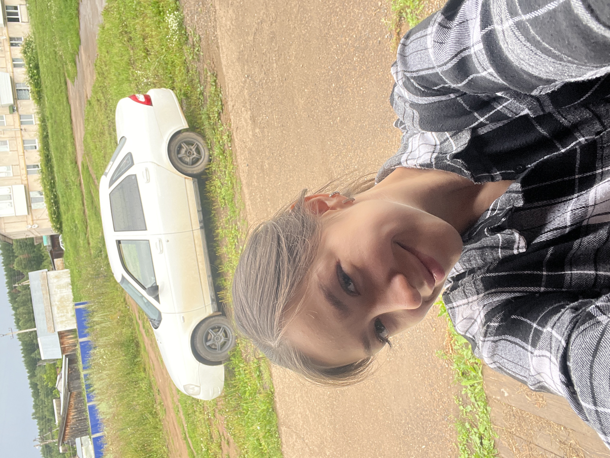 A young woman with brown hair pulled back, wearing a black and white plaid shirt, is taking a selfie in front of a white sedan. The car is parked on a dirt road next to a grassy area with some wildflowers. In the background, there are several buildings, including a multi-story apartment building and some smaller wooden structures, surrounded by trees. A power pole is visible to the left. The scene appears to be in a rural or semi-rural setting, likely in the city of Kemul, Russia, given the architecture and vegetation. The sky is overcast.