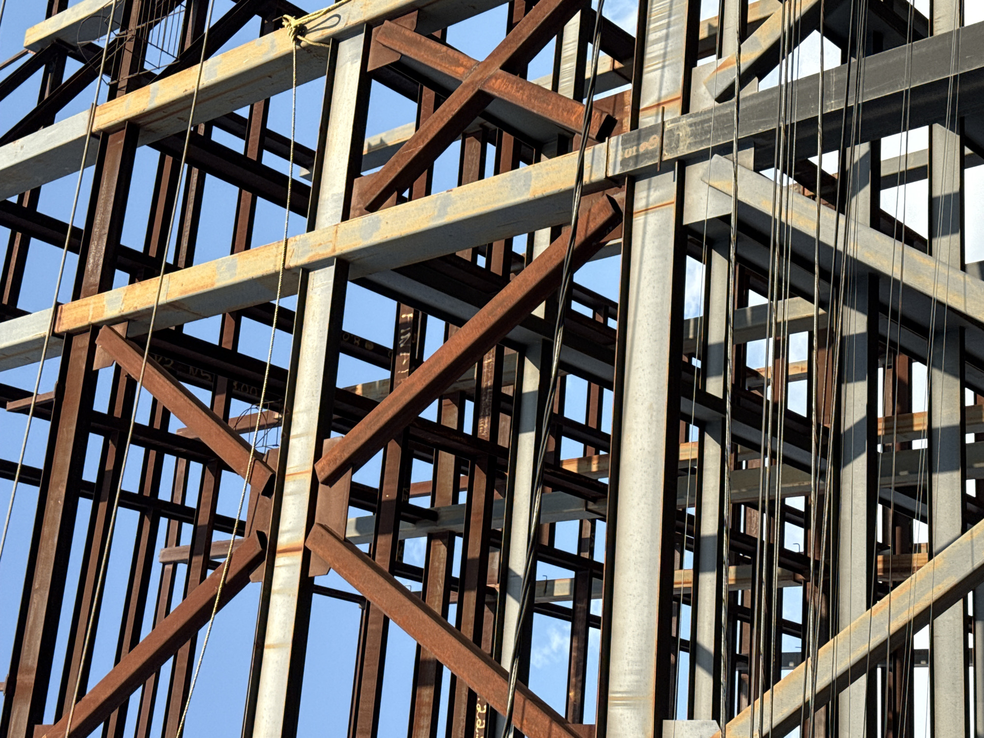 The image shows a steel framework under construction against a blue sky. The framework is composed of both gray and rust-colored steel beams. Some vertical cables are interspersed within the structure. The location is likely Puebla, Mexico, based on similar structures observed in that city.