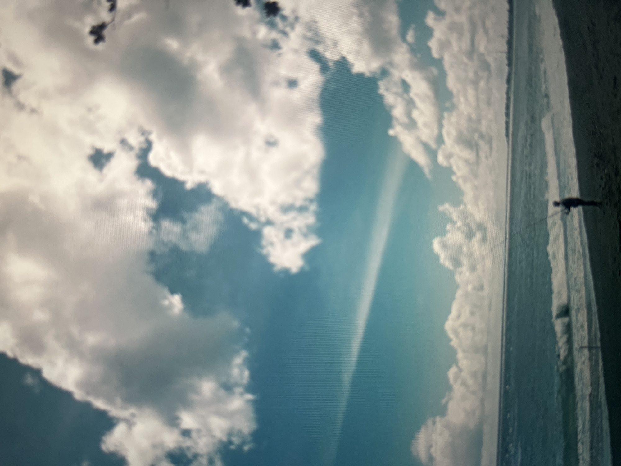 A lone figure stands on a dark sand beach, casting a fishing line into the choppy turquoise ocean under a dramatic sky filled with large, white cumulus clouds. The water exhibits moderate waves breaking on the shore. In the distance, a faint, light-colored line suggests a horizon or a distant landmass. The overall scene is a coastal landscape under a partly cloudy sky, with a single individual engaged in the activity of fishing. There are no discernible structures or specific landmarks visible that would definitively place the scene within Cirahayu, Indonesia, other than the provided contextual information.