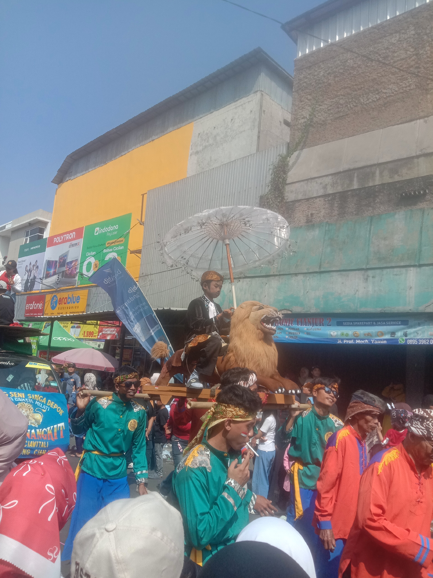 This is a medium shot of a street parade in Cianjur, Indonesia, under a clear blue sky. In the foreground, several men in traditional attire, primarily green shirts and blue pants, carry a large, light brown, faux lion effigy on a wooden platform. Some men are wearing headbands, and one is smoking a cigarette. Behind the lion, a young boy in a black outfit sits atop the effigy, holding a translucent white umbrella. The parade is passing by buildings with advertisements. One building has prominent yellow and orange facades with signage that includes "POLYTRON" and "Indodana." Another building displays a large blue banner with text that appears to be in Indonesian, and a phone number "0895 3962 0..." is visible. The environment suggests an urban setting with commercial activity.