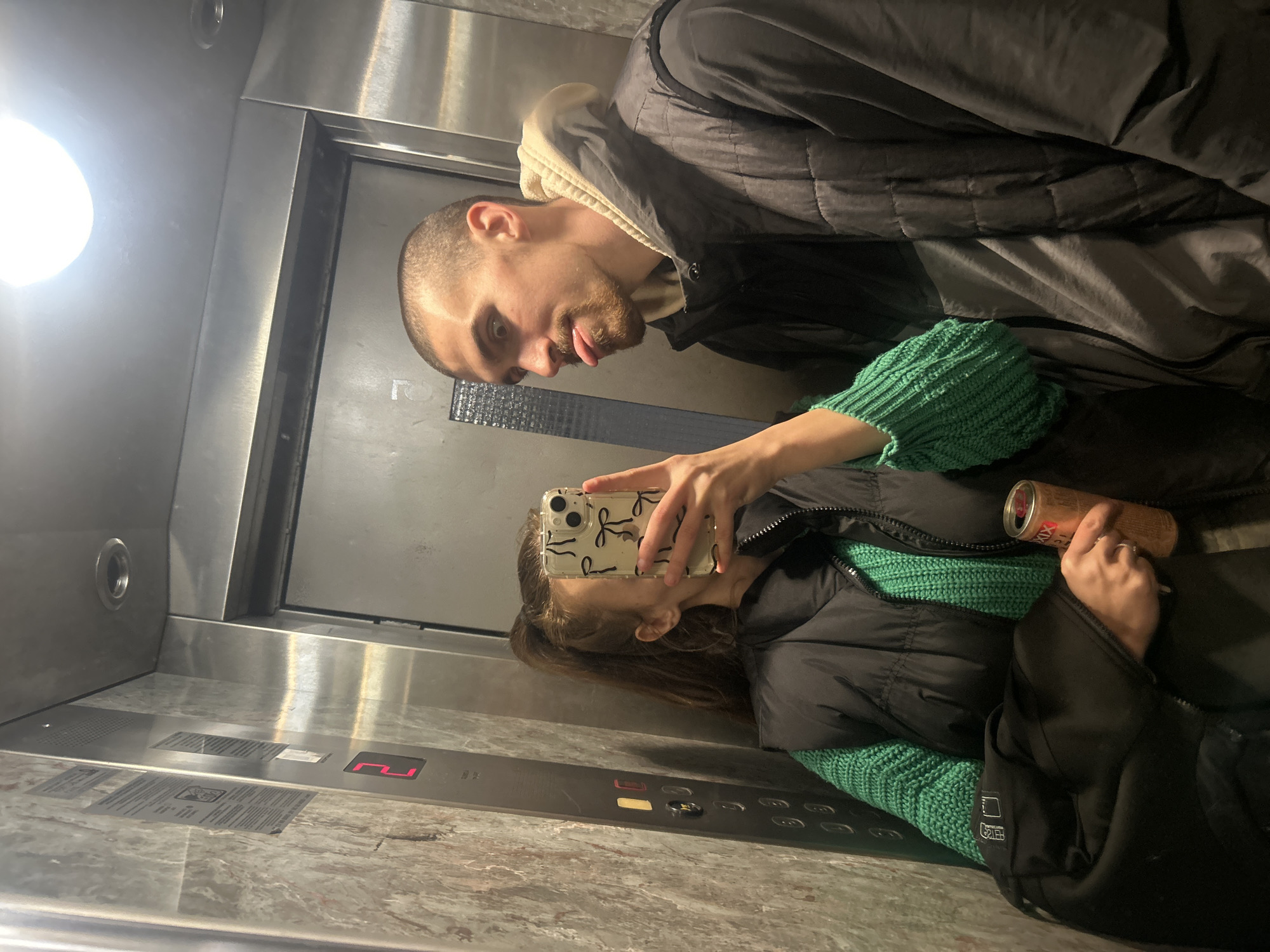 A selfie photograph inside an elevator car. A man with a shaved head, beard, and wearing a black jacket with a tan hoodie underneath, is visible. A woman, holding a cell phone with a case decorated with bows, is also visible in the reflection. She is wearing a black jacket and a green knitted sweater. She is also holding a beverage can. The elevator has stainless steel walls and a marble floor. The floor number indicator shows the digit "2" lit in red. The elevator buttons are visible. The location is Burgas, Bulgaria.