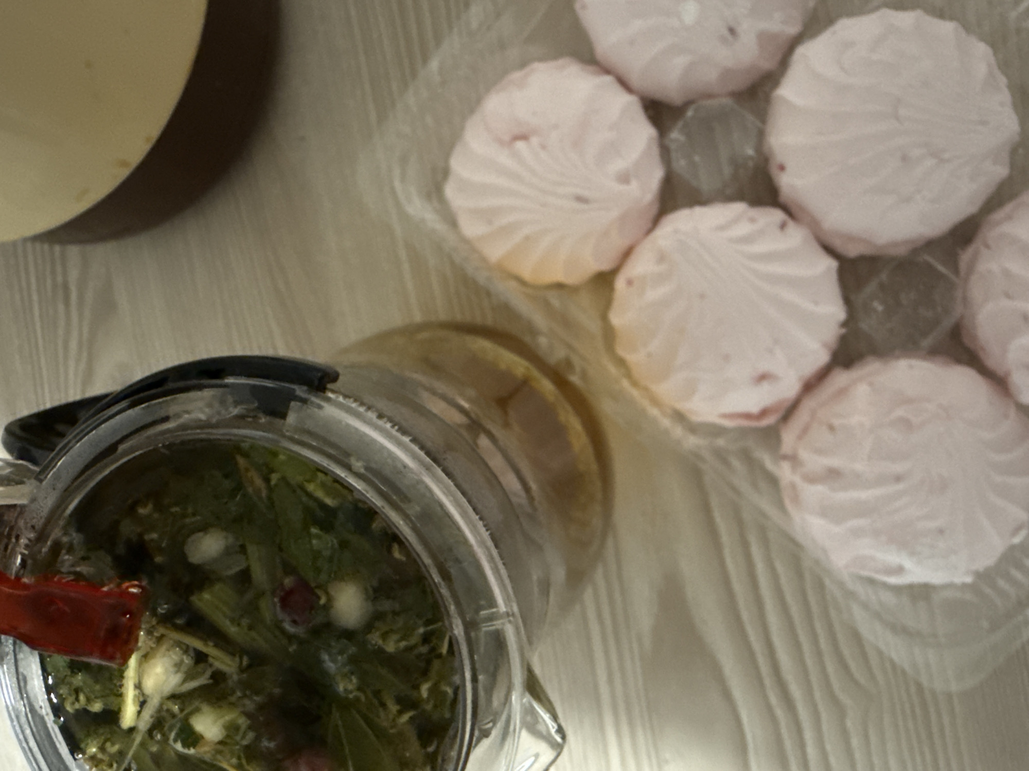 An overhead shot captures a cozy arrangement of tea and sweets on a light-colored, textured surface, possibly wood or laminate.
In the foreground and to the left, a clear glass teapot or infuser contains steeping herbal tea. The tea is a mix of green leaves, stems, small white buds or berries, and some dark red or purple elements, all immersed in light brown liquid. The teapot has a black handle and a visible red clip or spout component.
To the right, a clear plastic container holds several pink, circular marshmallows (zefir). Each marshmallow features a distinct swirled pattern on its top surface. The container suggests they are packaged confectionery.
Partially visible in the upper left corner is a section of a light-colored, possibly cream or beige, cup or bowl.
The lighting appears soft and even, suggesting an indoor setting. There are no people present, no visible text, and no specific indicators of time of day or weather. The scene evokes a sense of quiet enjoyment or a relaxing break.