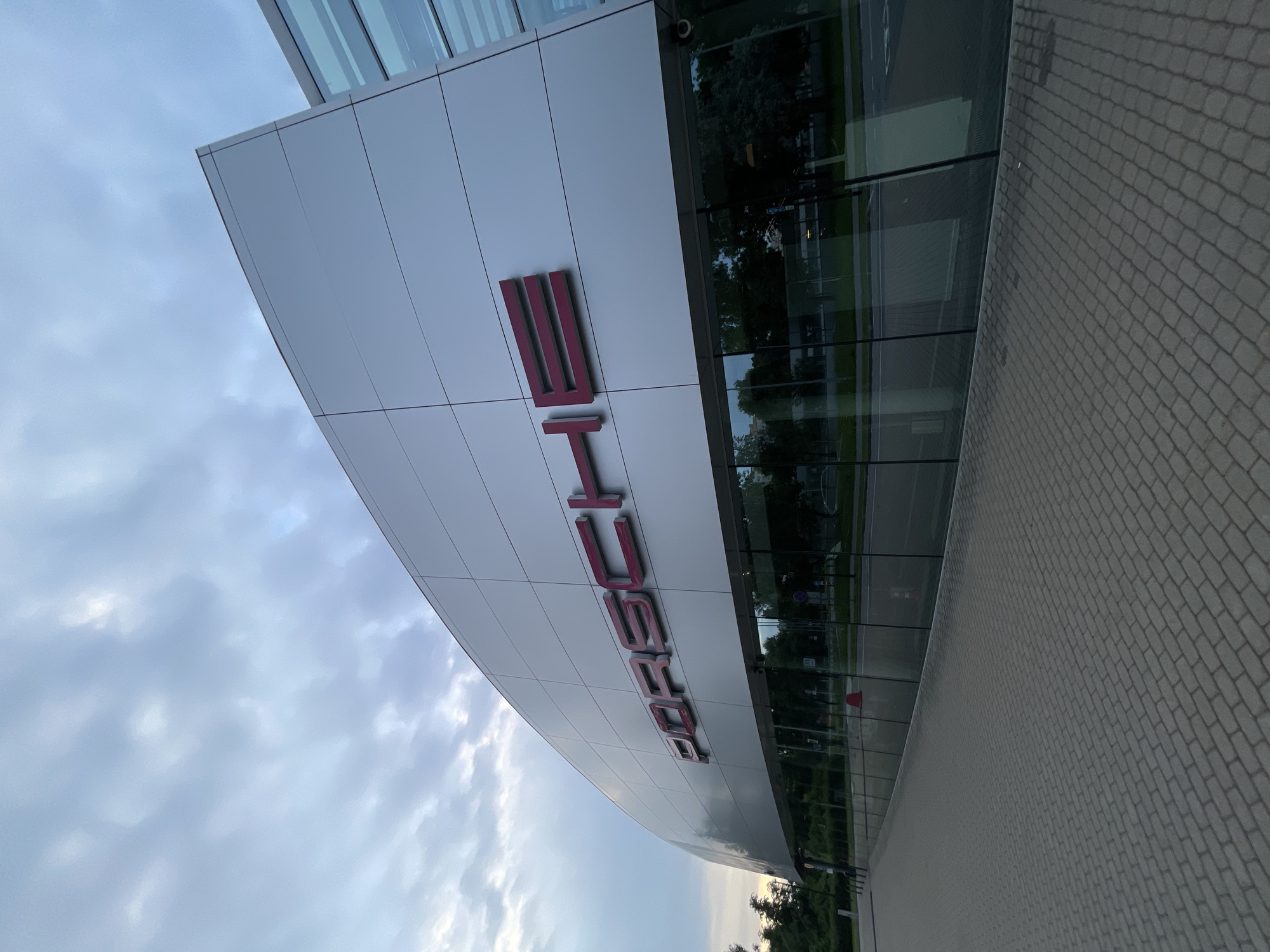 The image shows the exterior of a modern building with large glass windows and a white facade. Large, three-dimensional letters in a deep red color spell out "PORSCHE" on the white surface of the building. The building appears to be a car dealership or showroom.
To the right of the building, there is a paved walkway made of interlocking gray bricks. Beyond the walkway and through the glass facade, trees and a street are visible, suggesting an outdoor setting. Reflections of trees and lights can be seen in the glass. The sky above is cloudy and appears to be either early morning or late evening, given the soft, diffused light.
There are no people prominently featured in the image, though a faint shadow of a person can be seen near the bottom left corner of the paved walkway. The overall scene conveys a sense of quiet and perhaps anticipation, as is often associated with car showrooms. The architectural style is sleek and contemporary. The visible text is the brand name "PORSCHE".