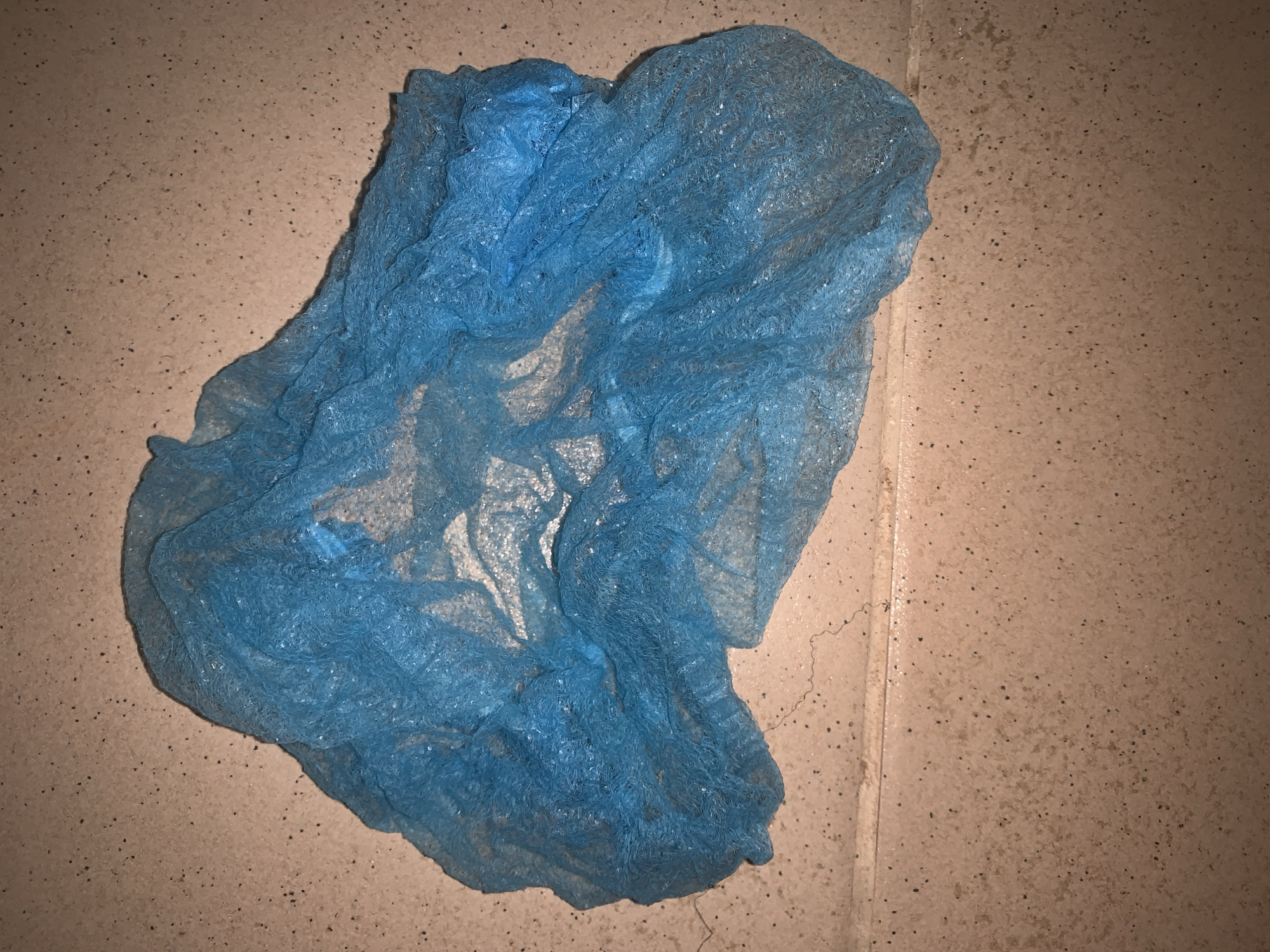 A crumpled, blue, disposable hair net lies on a tiled floor. The tiles are beige with small black speckles. The hair net is made of a thin, non-woven material and has an elasticated edge. The lighting in the image is dim, casting shadows. There is no indication of the location within a city.