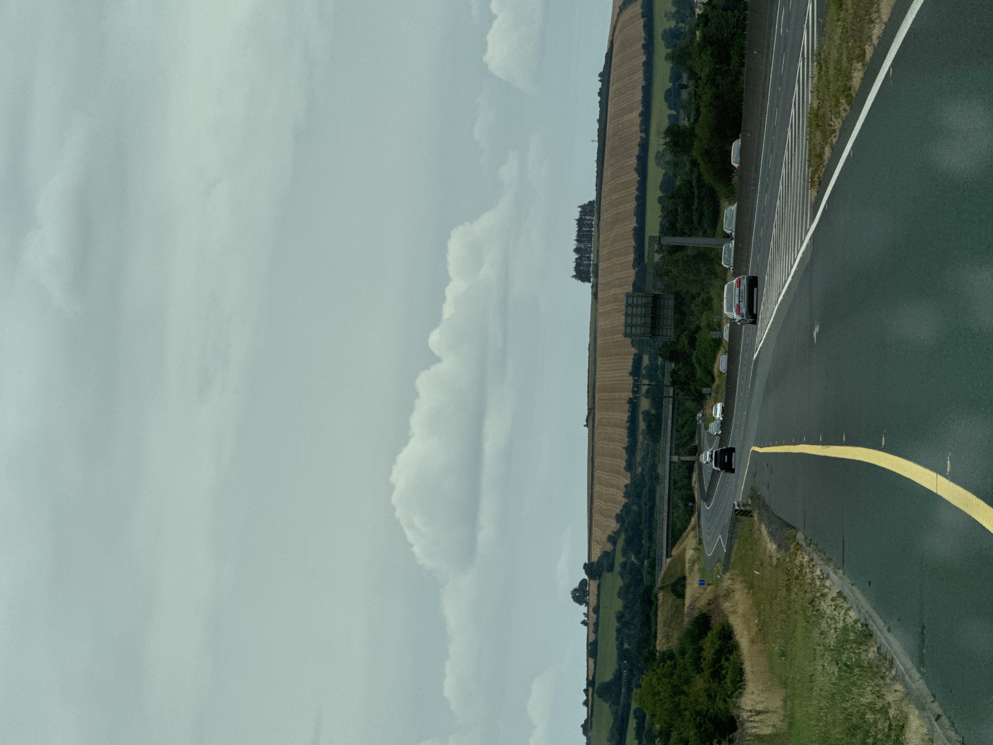 An eye-level, medium shot captures a view of a multilane highway curving through a rural landscape under an overcast sky in Hacklow, Ireland. The road, paved with dark asphalt, is divided by a bright yellow line on the left and a white line on the right, with a series of horizontal white lines marking a transition in the road on the far right. Several vehicles, including a black SUV and a silver sedan, are visible on the highway, with others in the distance. The road is bordered by grassy verges and shrubs. In the background, rolling hills are covered with a patchwork of green fields and a harvested golden field, with a cluster of trees on a distant ridge. A concrete overpass is visible spanning the highway in the mid-ground, and a tall, square sign structure stands to the left of the road.