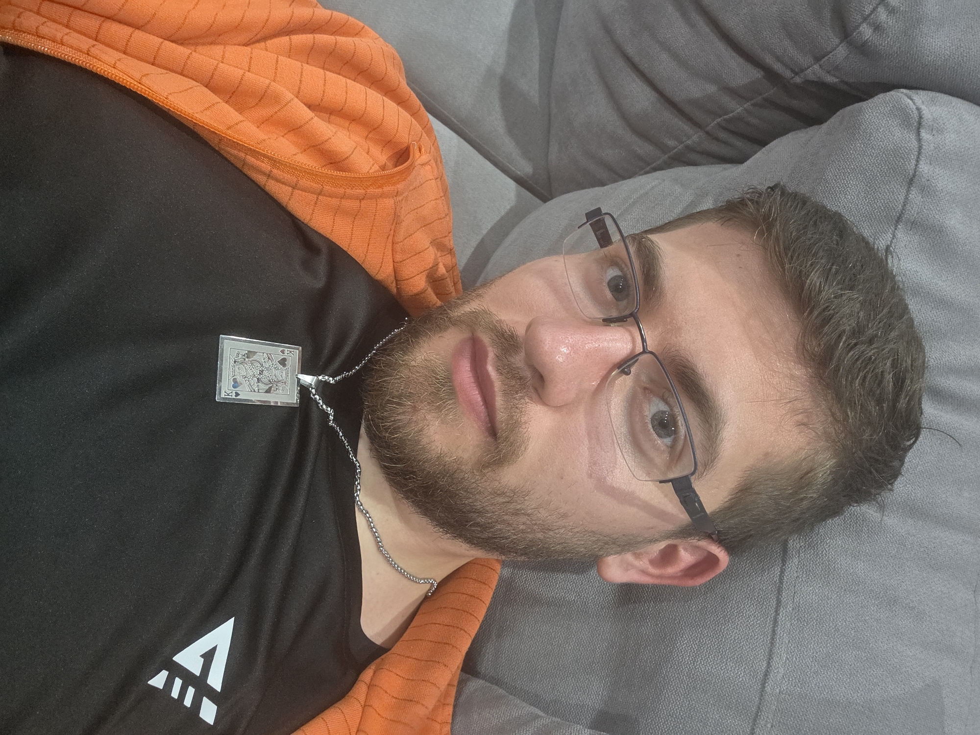 A man with short brown hair and a beard, wearing glasses, a black t-shirt with a white logo, and an orange jacket, is lying on a grey couch. He is wearing a silver chain with a pendant depicting a playing card, likely the King of Spades. No specific information indicates that this image was taken in Yagodovo, Bulgaria. The location is unknown.