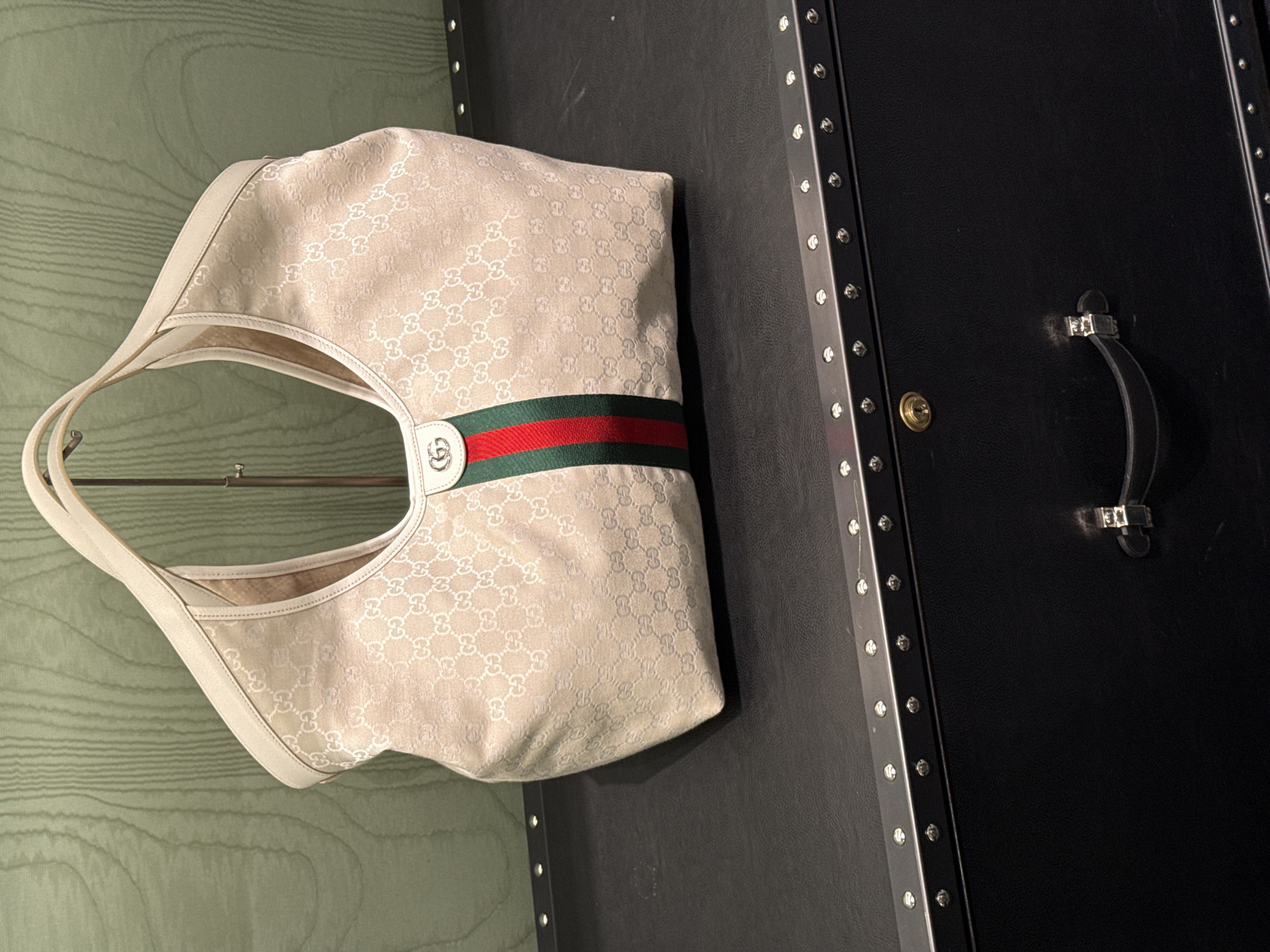 The image shows a Gucci handbag resting on a black leather trunk. The handbag is light beige with a repeated "GG" pattern, a green and red striped band, and a white leather handle. The trunk has a black leather exterior, metal corners and trim with rivets, and a black leather handle with metal hardware. The background is a light green wall with a textured surface. The location and the city of Puebla, Mexico are not inferable from the image.