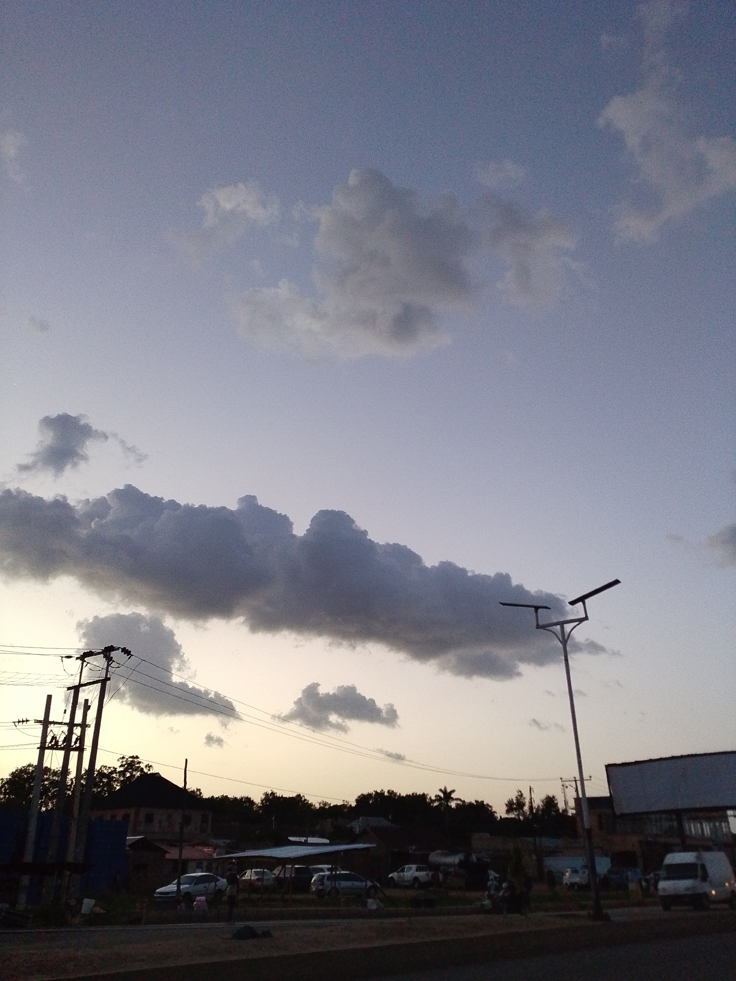 Captured in Kuru Dungus, Nigeria, the photograph shows a street scene at dusk. The sky is a pale gradient of blue and grey, dotted with scattered clouds. Overhead power lines and a street lamp intersect the skyline. The horizon is lined with trees and buildings. Cars are parked along the street. A white van is prominent on the right, and individuals are present along the roadside. The foreground is a road surface.