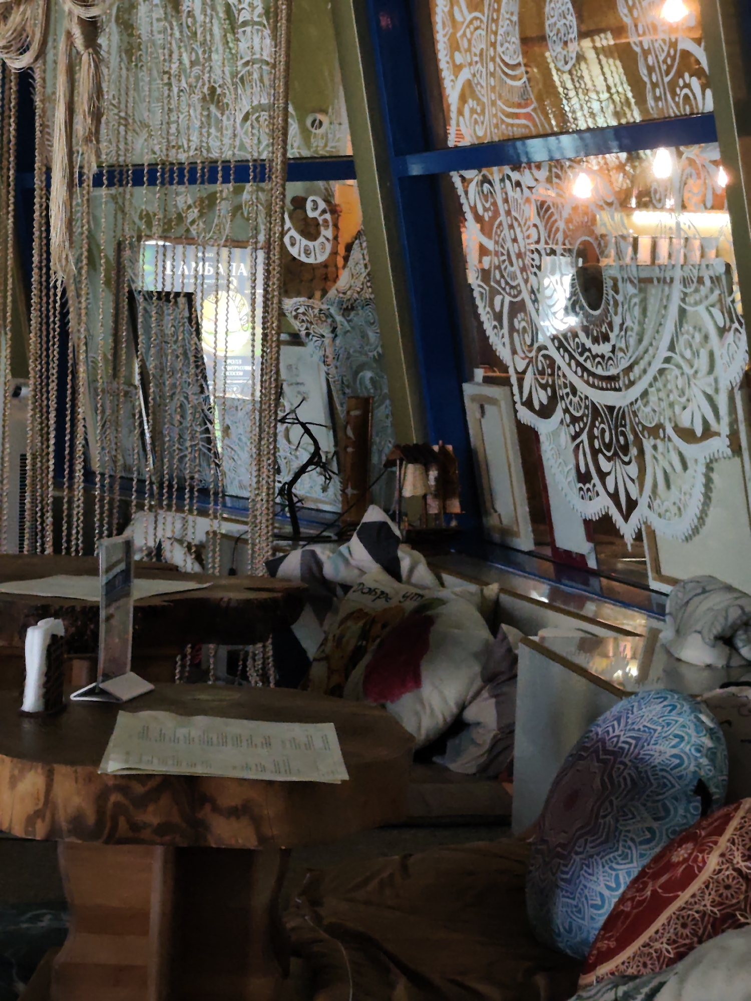 This image depicts the interior of a cafe in Sochi, Russia. The scene is characterized by a bohemian, eclectic decor. A round, wooden table is in the foreground, with a menu or document resting on its surface. To the left, a napkin dispenser and a menu holder are visible. Behind the table, a beaded curtain hangs, separating areas of the cafe. The background features large windows with intricate white patterns, possibly stencils or decals, resembling mandala designs. Through the windows, reflections show lighting and possibly more of the cafe's interior. The seating area is furnished with cushions displaying various patterns, including one with a prominent blue mandala design. A decorative branch is placed near the window. The overall ambiance suggests a relaxed and artistic atmosphere within a commercial establishment.